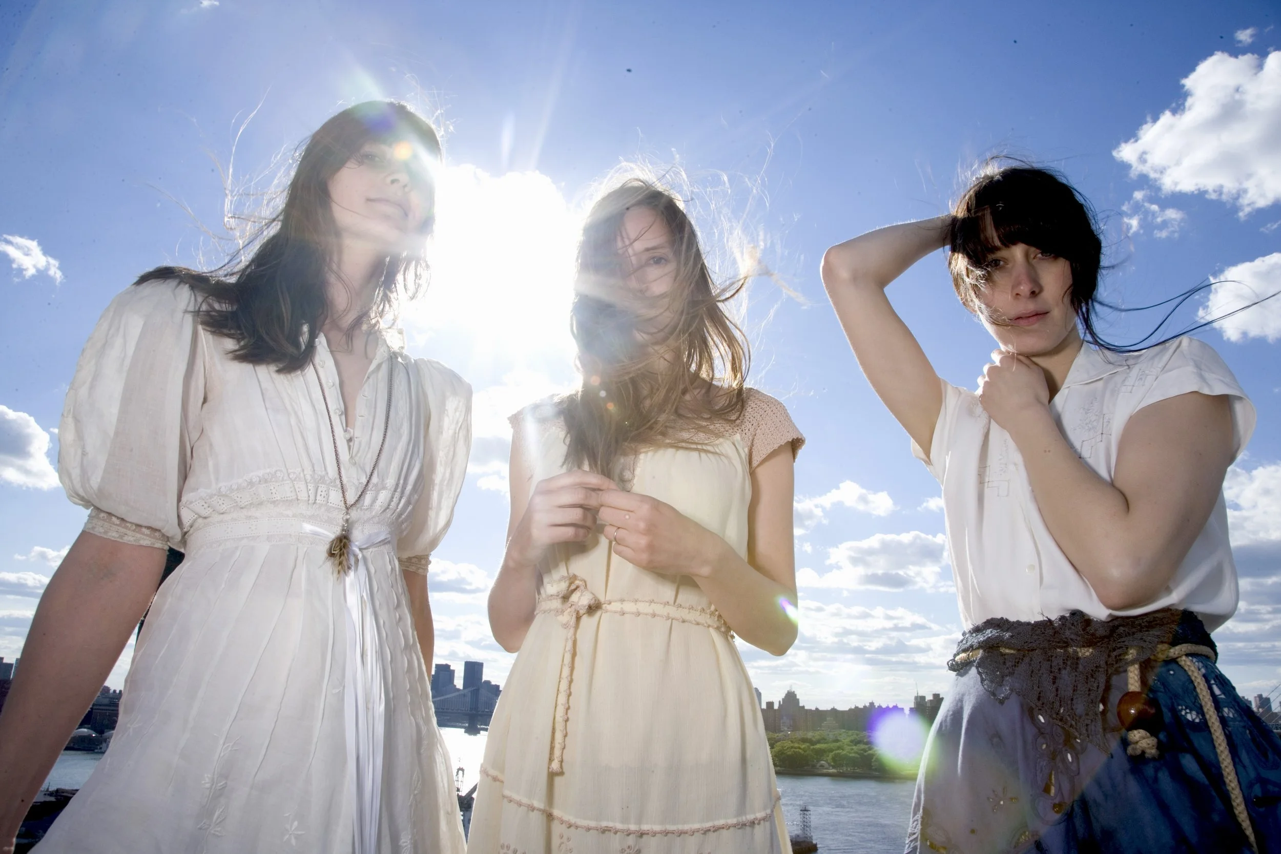 Drei junge Frauen stehen im Freien bei strahlendem Sonnenschein vor einer Wasserlandschaft mit Stadt im Hintergrund, tragen leichte Sommerkleidung und schauen in die Kamera.