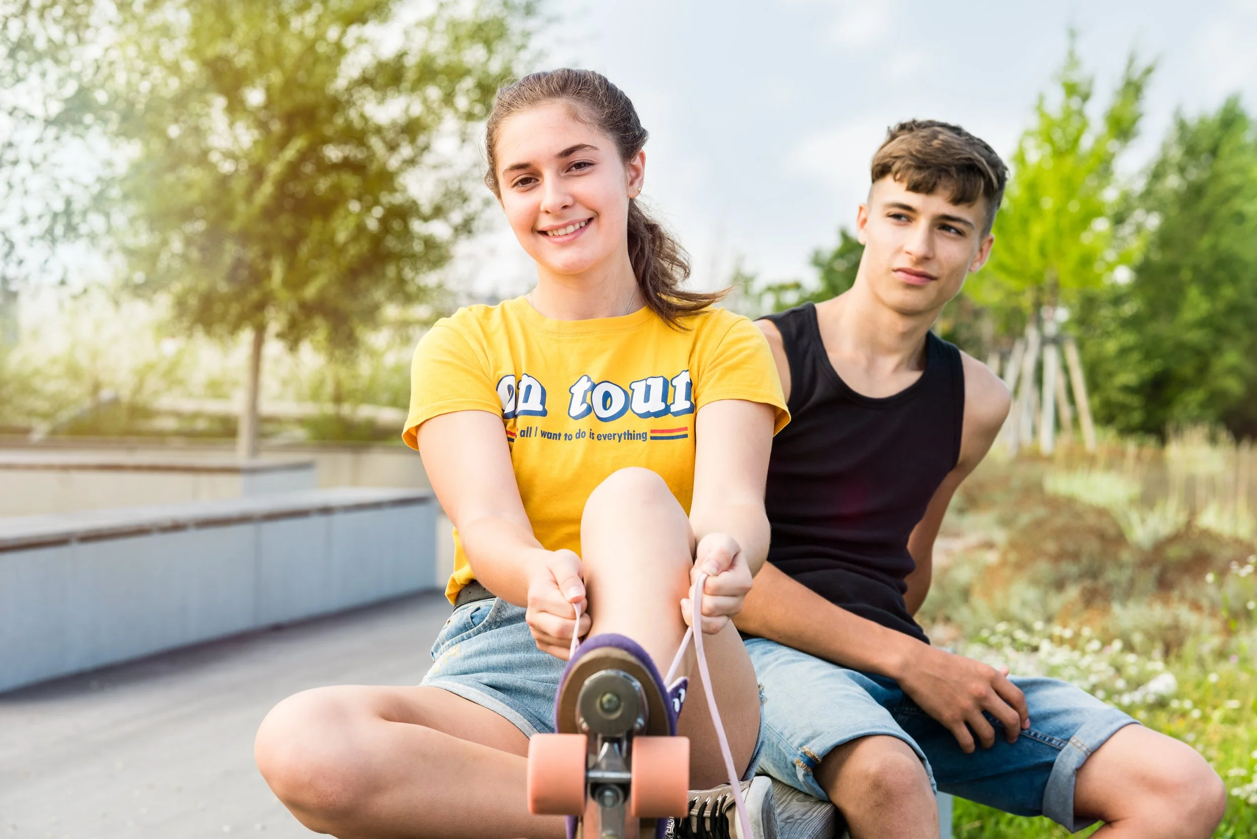 Zwei Jugendliche sitzen im Park, das Mädchen beim Entspannen auf einem Skateboard, während der Junge neben ihr sitzt. Es ist sonnig, und im Hintergrund sind Bäume und eine grüne Parklandschaft sichtbar.