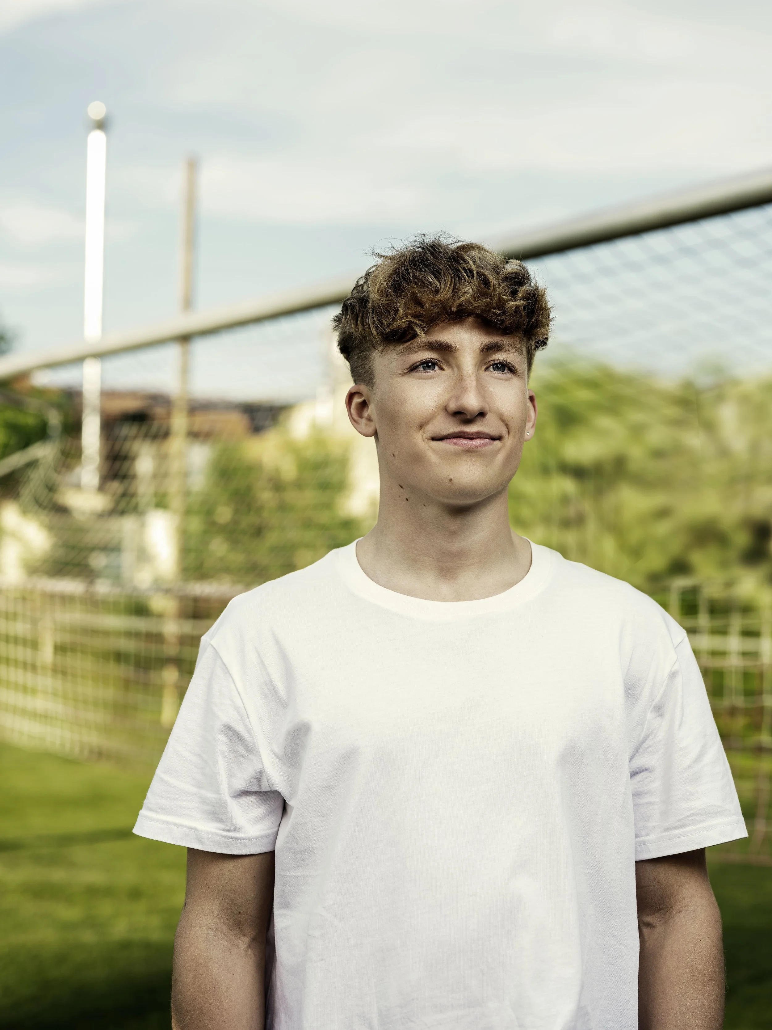 Junge steht vor einem Fußballtor auf dem Sportplatz, trägt ein weißes T-Shirt, lächelt und schaut in die Kamera, im Hintergrund Bäume und ein blauer Himmel.