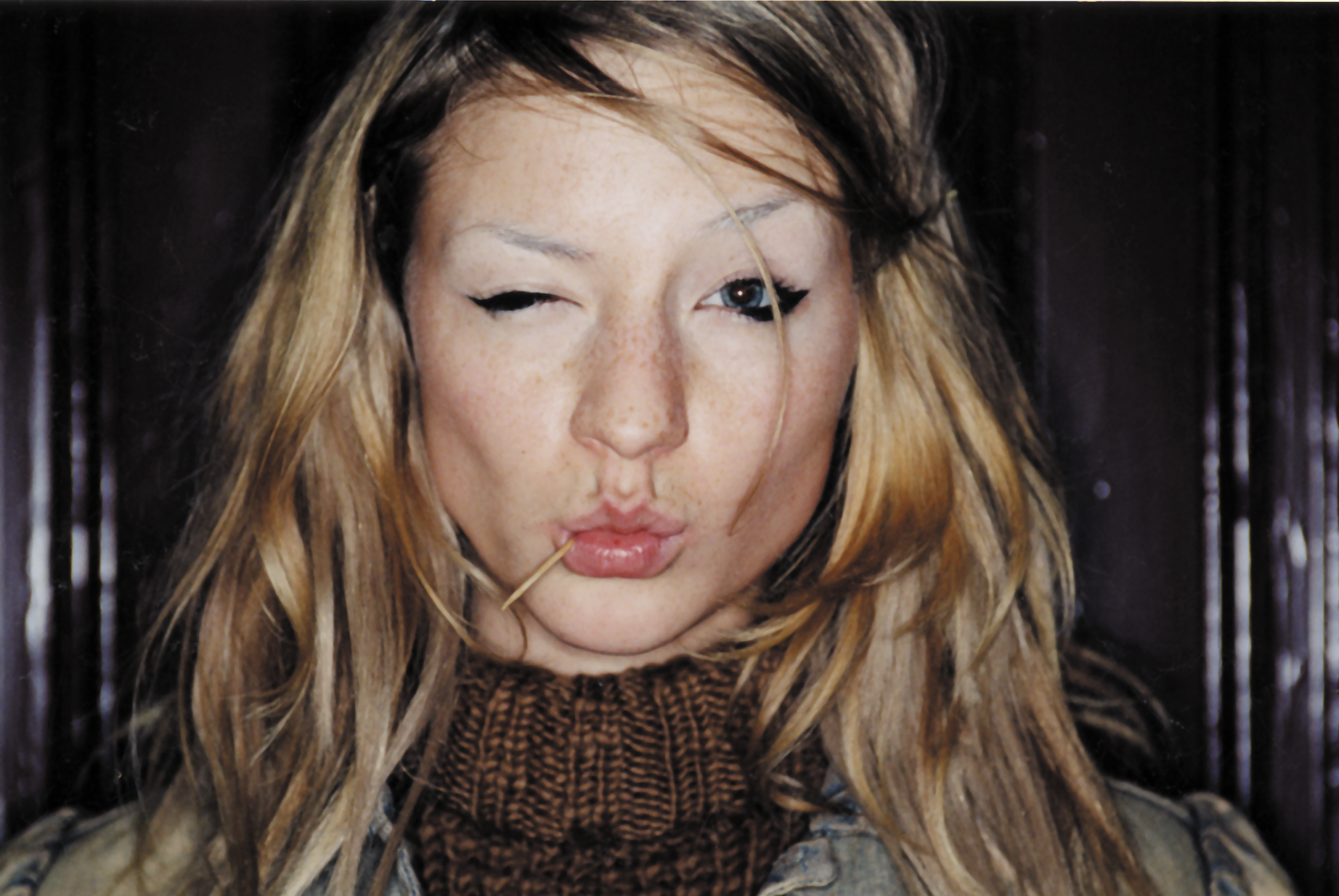 Junge Frau mit langem, blondem Haar, eine Augenpartie spielt sie mit Wimperntusche und hat ein kariertes Hemd an, mit einem Zahnstocher im Mund, vor dunklem Hintergrund.