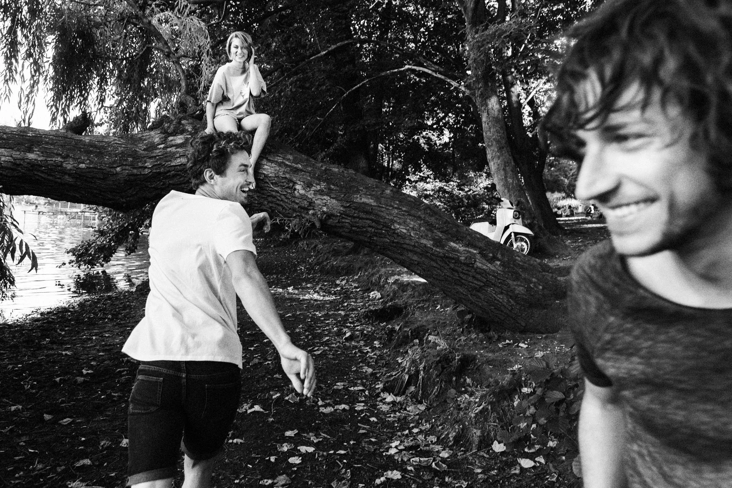 Drei Jugendliche lachen und spielen im Park, einer sitzt auf einem Baum, die anderen stehen am Boden. Es ist schwarz-weiß, die Umgebung ist voller Bäume und Blätter.