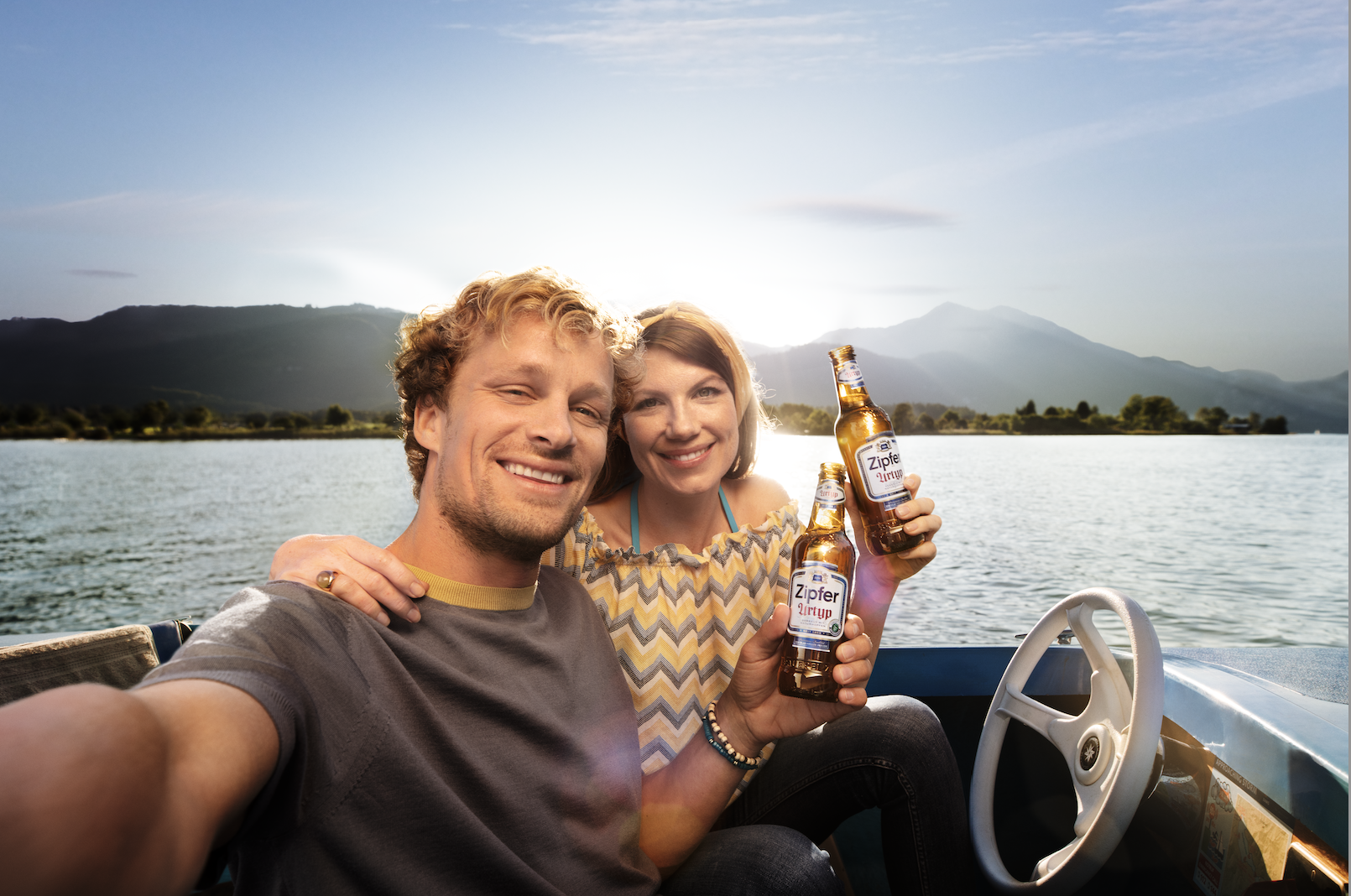 Ein lachendes Paar macht ein Selfie auf einem Boot mit einem See, Bergen im Hintergrund, während sie Bier trinken.