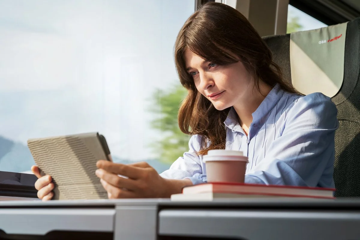 Frau mit braunen Haaren liest ein Buch auf einem Zug, mit einem Kaffeebecher vor sich.