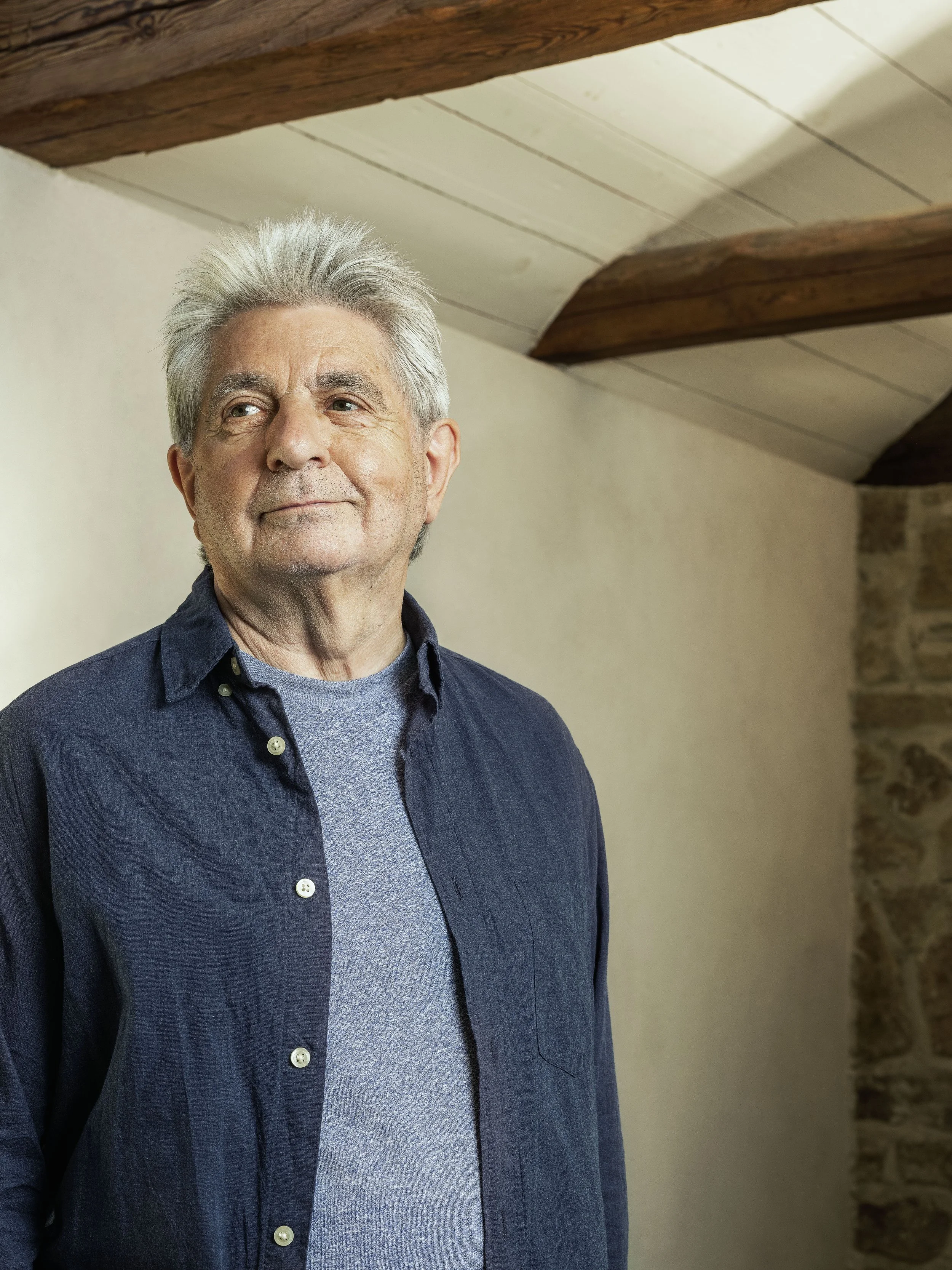 Ein älterer Mann mit grauem Haar, trägt ein dunkelblaues Hemd über einem grauen T-Shirt, steht in einem Raum mit Holzbalkendecke und einer Steinmauer im Hintergrund.