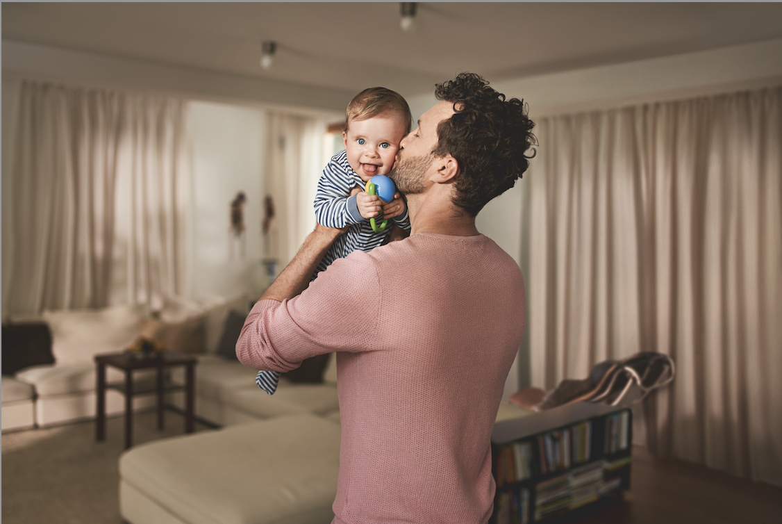 Vater hält lachendes Baby in einem Wohnzimmer mit gemütlicher Einrichtung und Vorhängen.