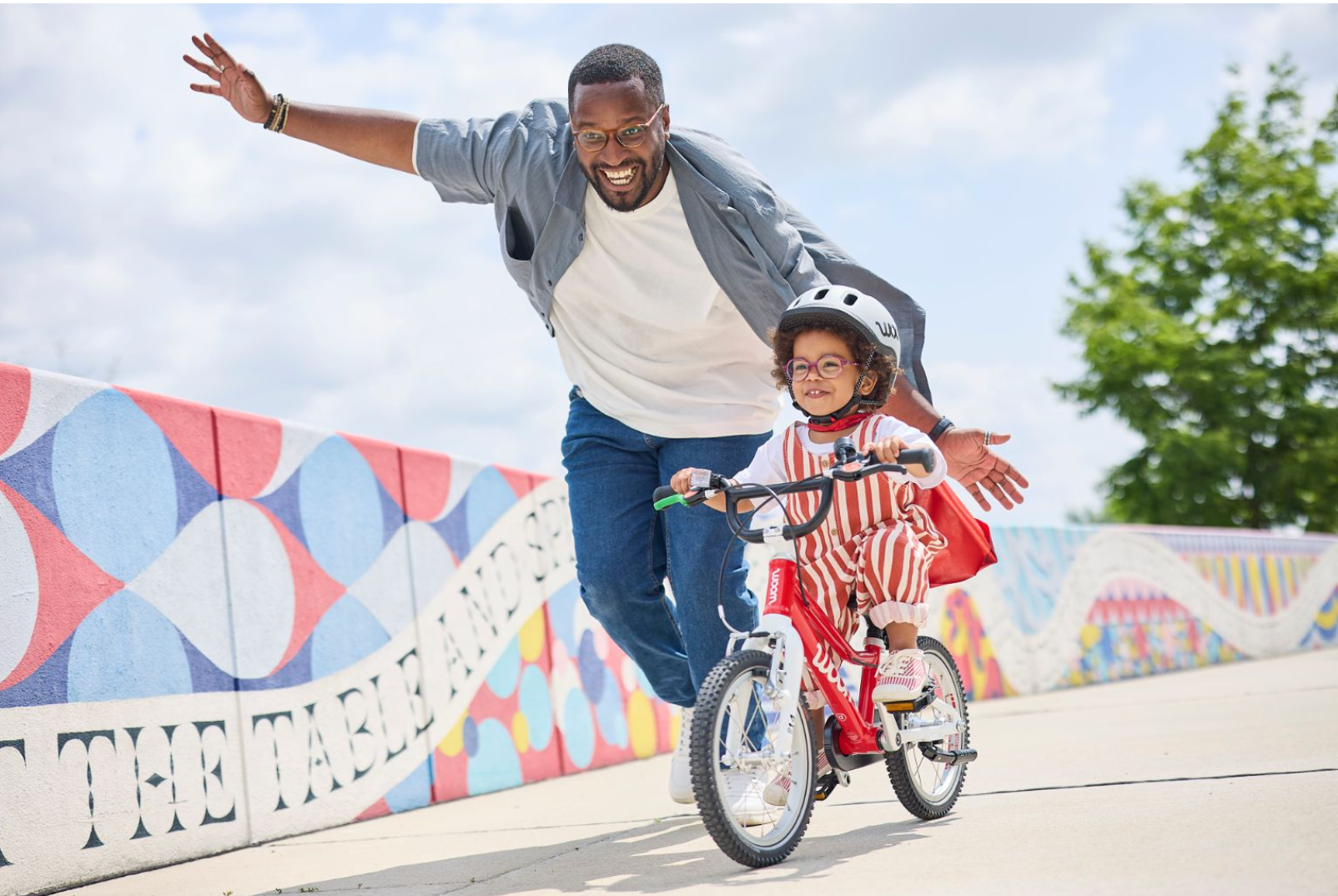 Ein Mann läuft lachend neben einem kleinen Mädchen, das Fahrrad fährt, während sie auf einer Brücke mit buntem Murmelnmuster radelt, bei sonnigem Himmel.