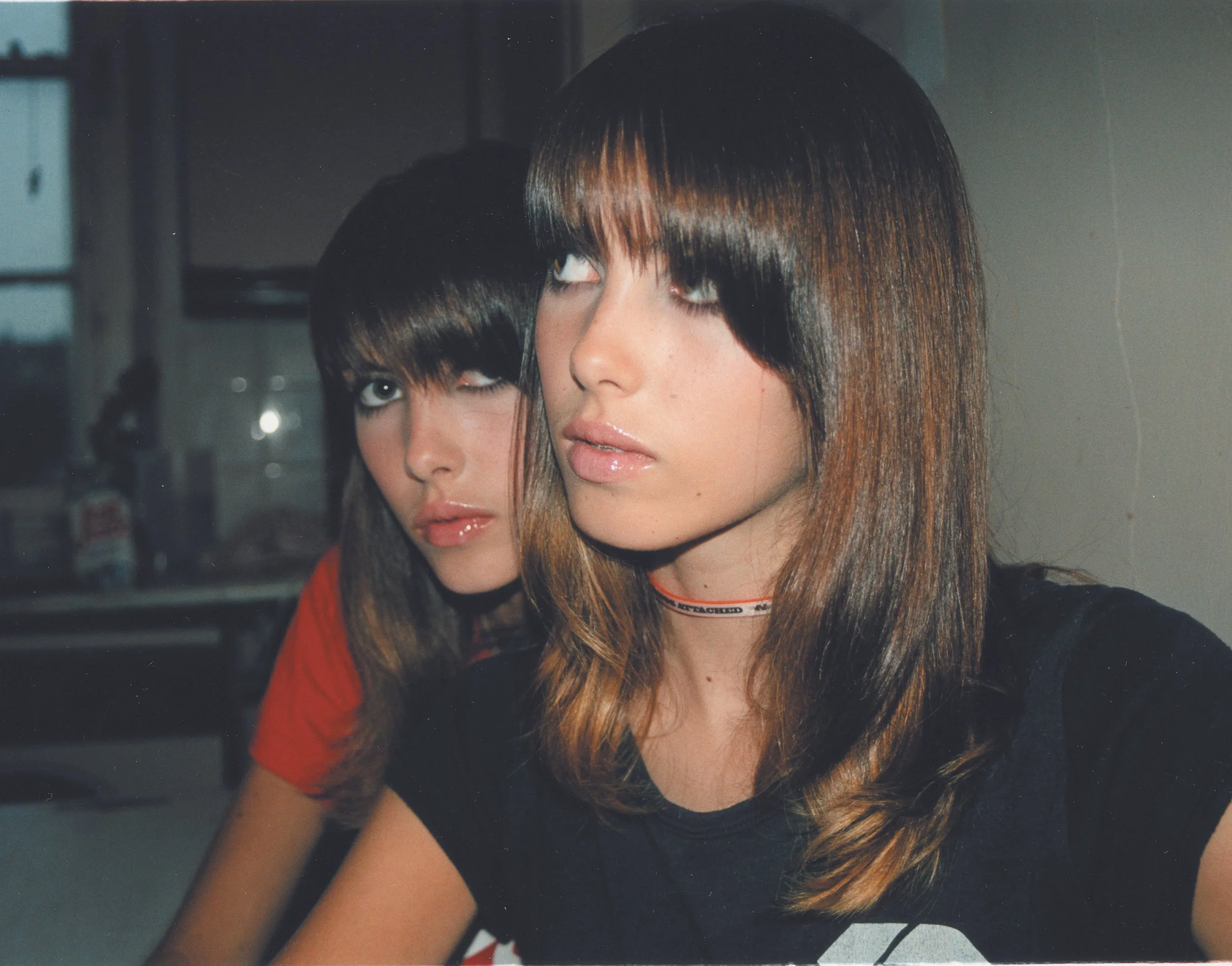 Zwei junge Frauen mit ähnlichen dunklen Haaren und welligem Haarschnitt posieren nebeneinander vor einem Fenster.