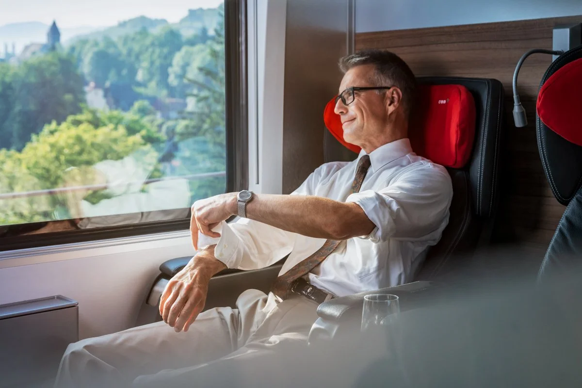 Ein Mann sitzt entspannt in einem Zug, lehnt sich zurück und schaut zufrieden aus dem Fenster auf die grüne Landschaft.