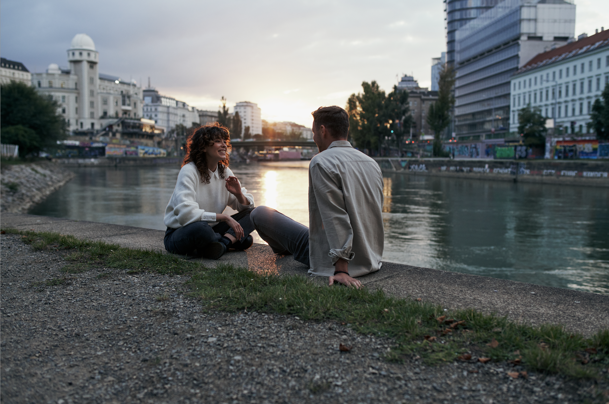 Zwei Menschen sitzen am Flussufer, unterhalten sich bei Sonnenuntergang, umgeben von modernen Gebäuden in einer Stadt.