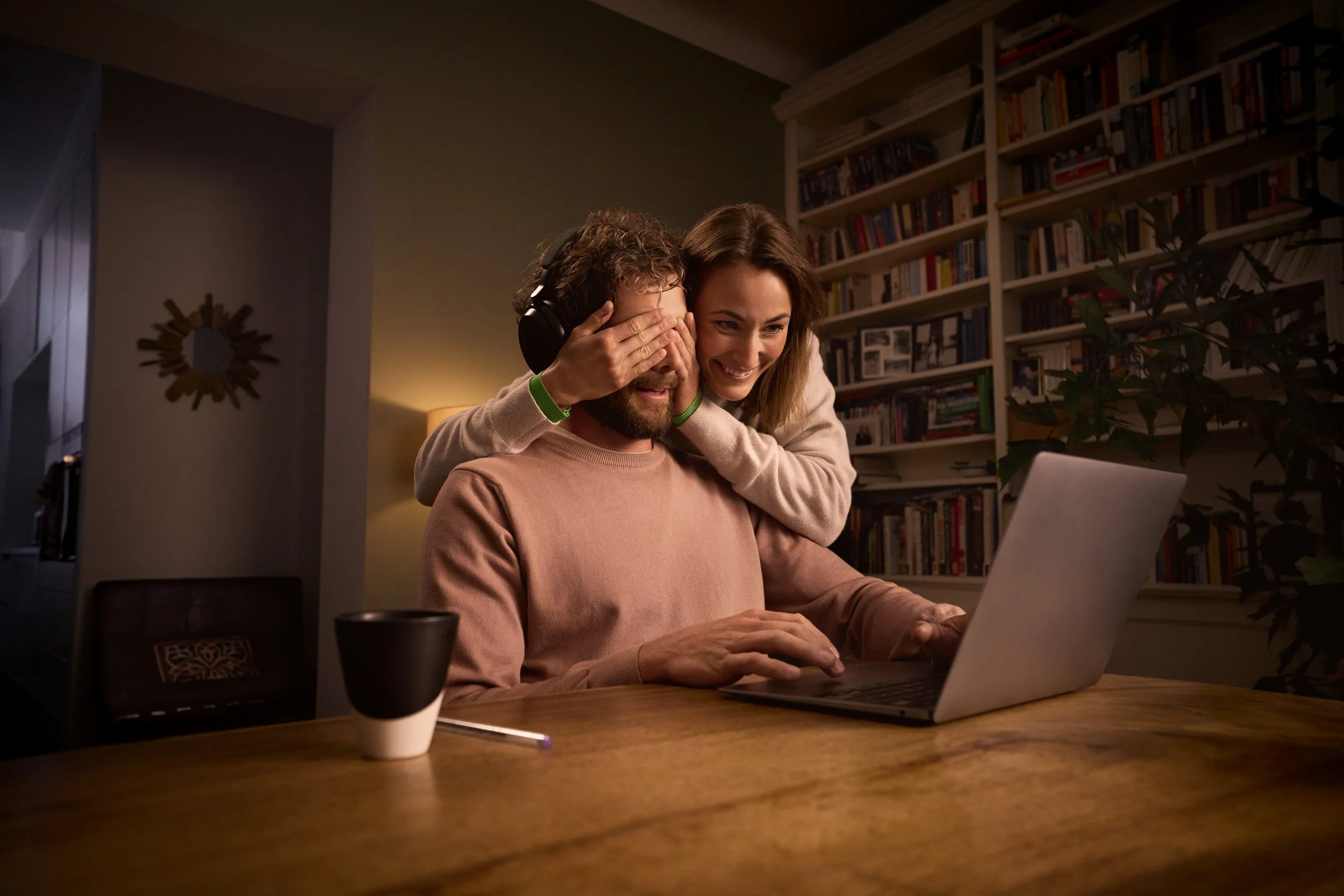 Ein lachendes Paar sitzt vor einem Laptop in einem Wohnzimmer, während die Frau dem Mann mit verbundenen Augen eine Überraschung bereitet.