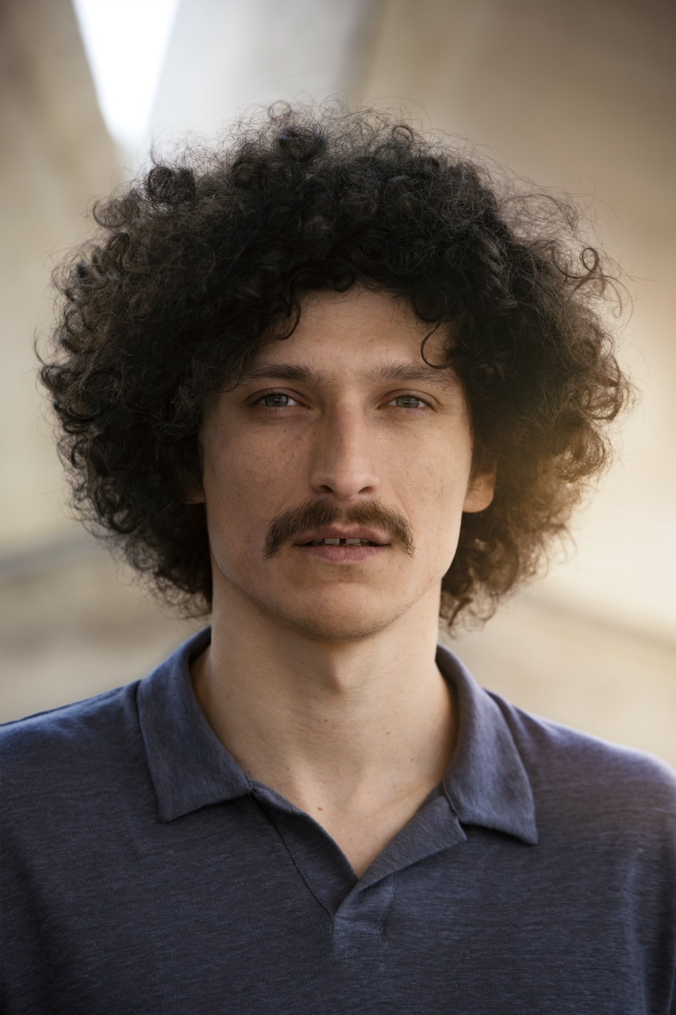 Junger Mann mit lockigem Haar, Schnurrbart und grauem Polo-Shirt, steht vor einem unscharfen outdoor-Hintergrund.