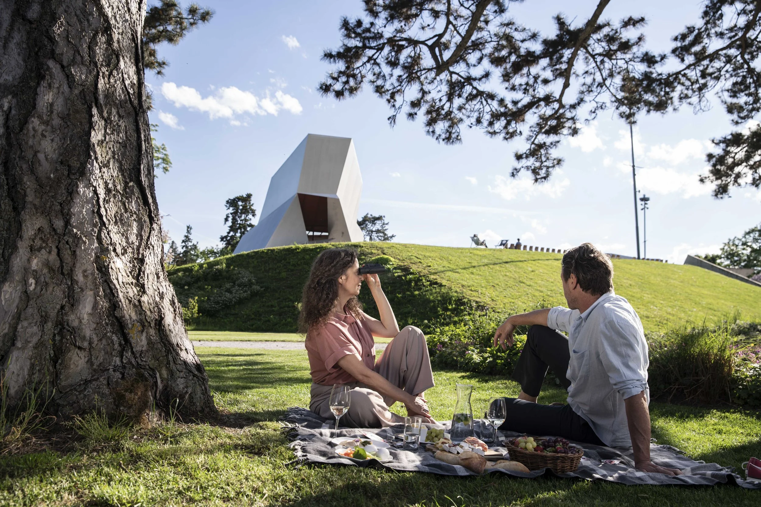 Zwei Menschen sitzen auf einer Decke im Park, es gibt Weingläser, eine Flasche Wasser, Obst und eine Karaffe, während sie sich unterhalten, im Hintergrund ein moderner Bau auf einem Hügel und Bäume unter blauem Himmel.