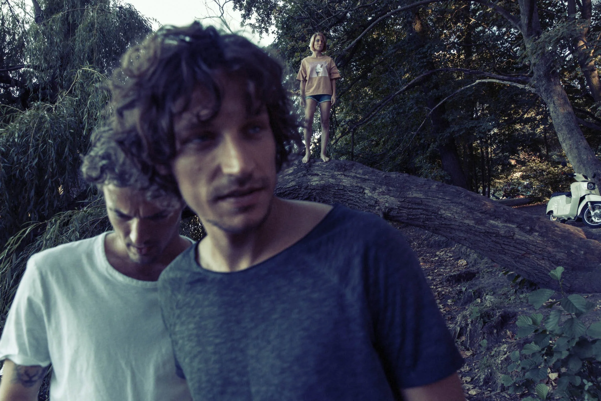 Zwei junge Männer im Vordergrund, einer schaut nach rechts, der andere nach unten, im Hintergrund liegt ein Baum, an dem ein Mädchen steht, und ein weißes Moped ist zu sehen, alles in einem parkähnlichen Bereich bei Dämmerung.