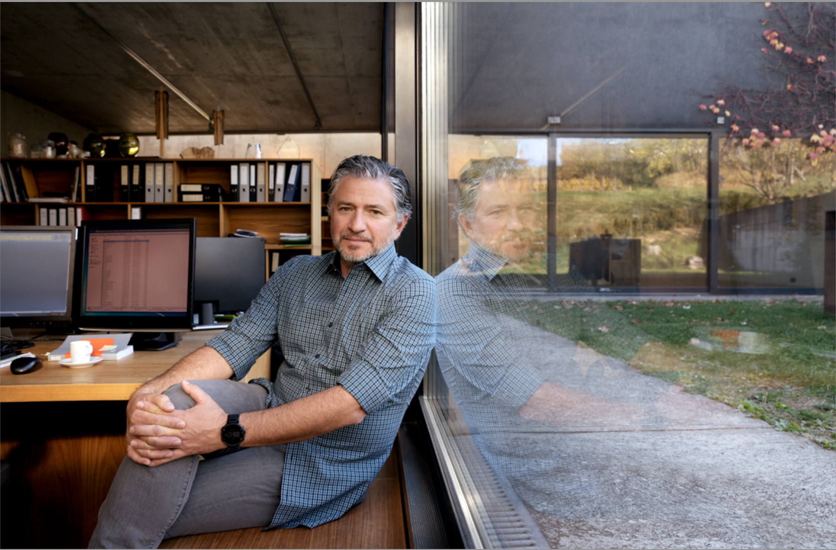 Mann sitzt in einem Büro neben einem großen Fenster mit Blick nach draußen auf eine grüne Landschaft, seine Reflexion ist im Fenster zu sehen.