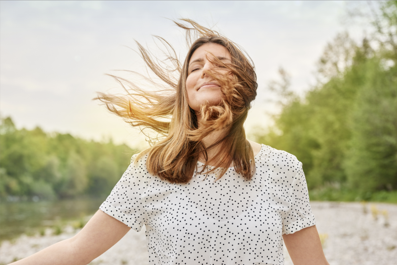 Eine glückliche junge Frau in einem weißen Oberteil mit schwarzen Punkten steht im Freien bei sonnigem Wetter. Ihre Haare wehen im Wind, während sie entspannt und zufrieden ist, umgeben von grünen Bäumen und Wasser im Hintergrund.
