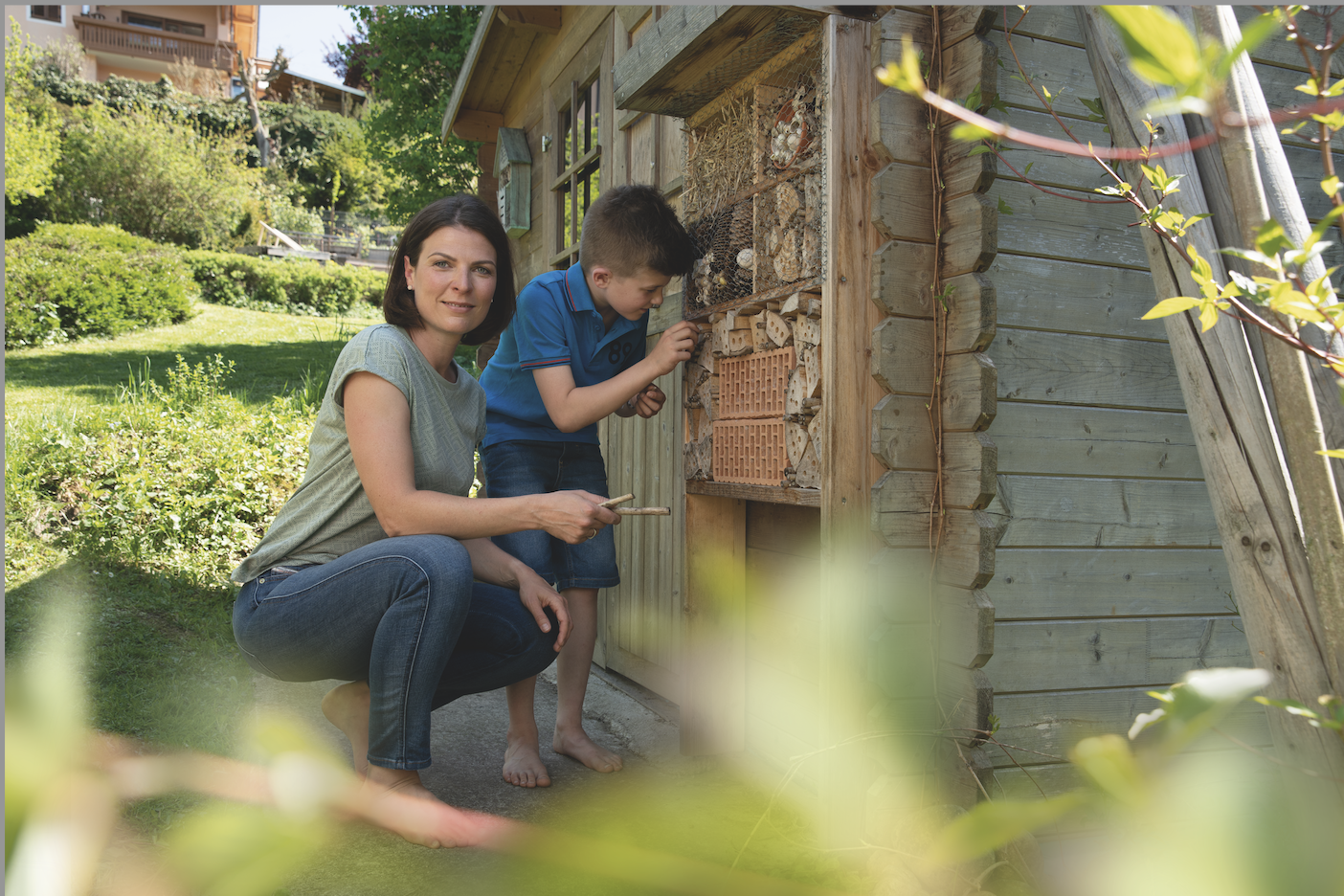 Eine Frau und ein Junge beobachten ein Insektenhotel an einem Gartenhaus. Es ist sonnig, und der Garten ist grün und gut gepflegt.