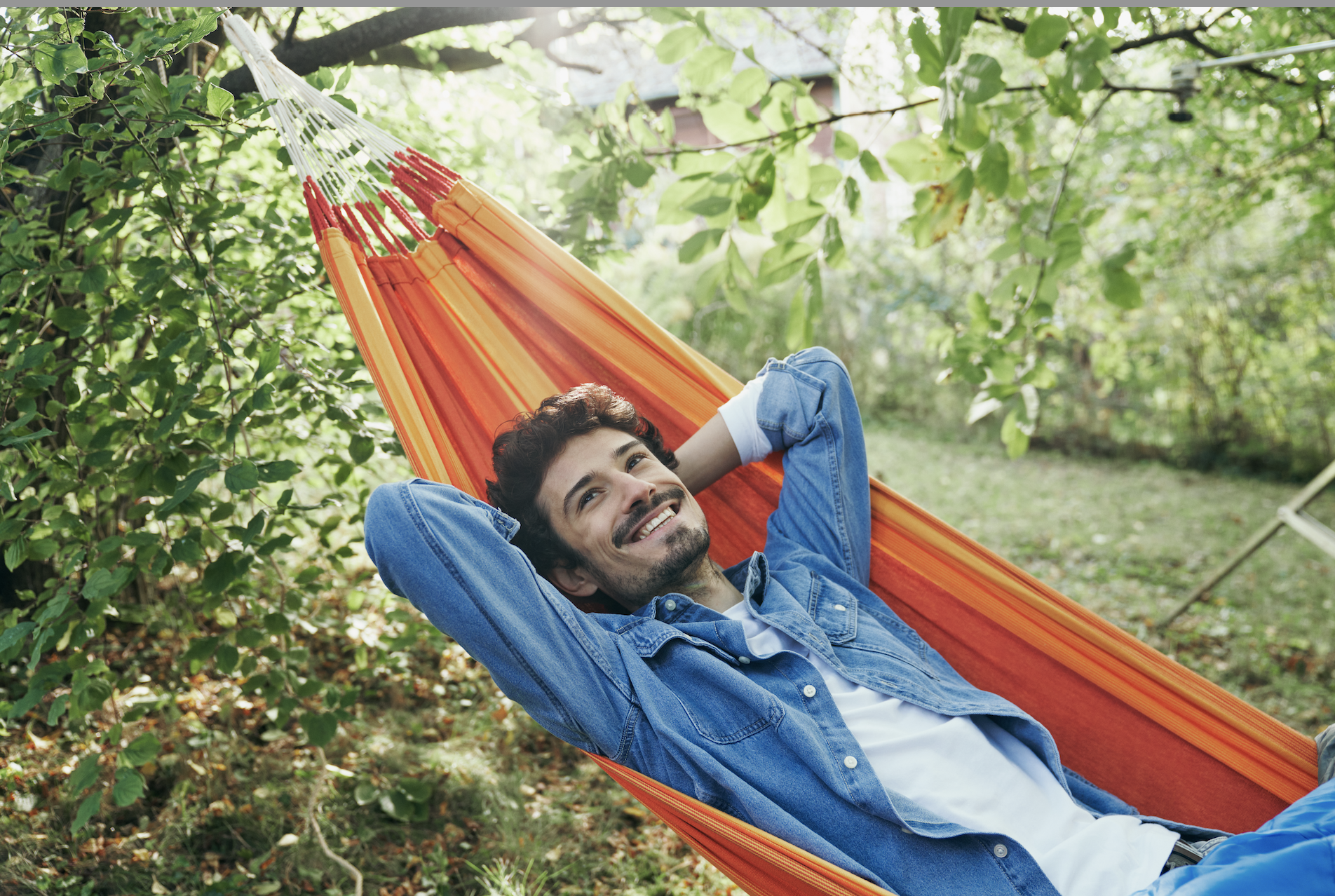 Ein entspannter Mann liegt in einer bunten Hängematte im Garten, umgeben von grünen Bäumen und Sträuchern, und lächelt glücklich.