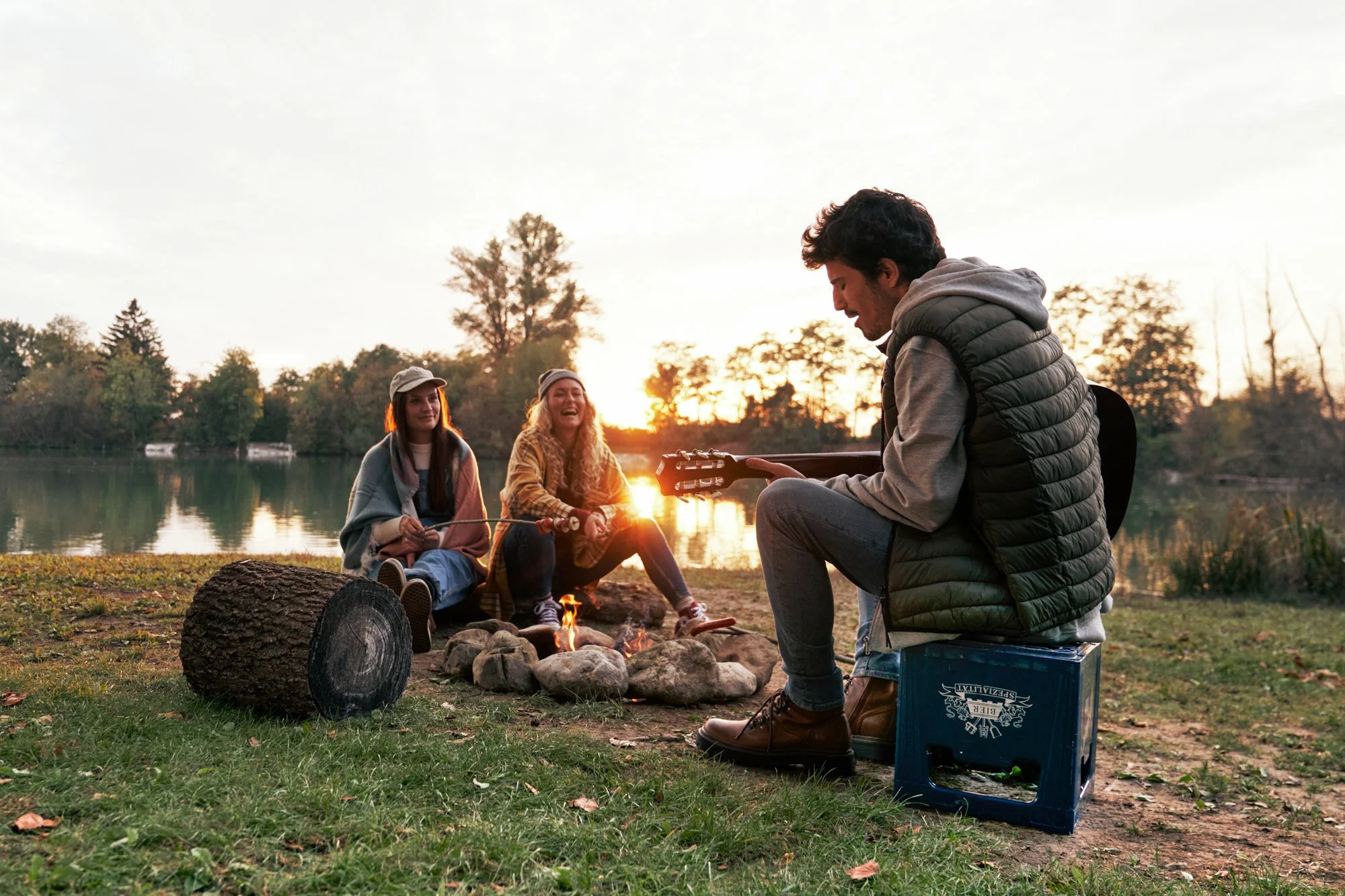 Gruppe von jungen Leuten sitzt am Lagerfeuer bei Sonnenuntergang, einige spielen Gitarre und lachen, im Hintergrund ein See und Bäume.