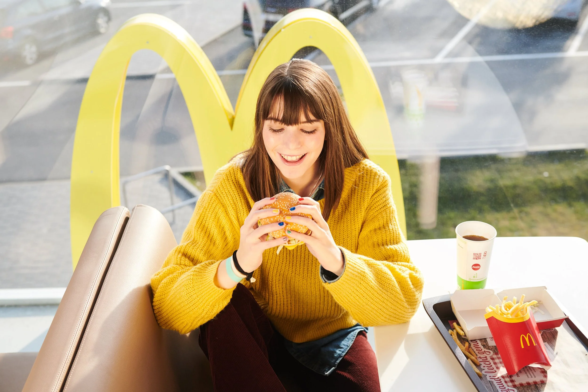 Junge Frau in einem McDonald's Restaurant, die einen Hamburger hält und lächelt, mit McDonald's Pommes und Getränk auf dem Tisch.