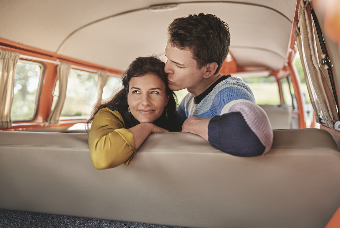 Ein junges Paar kuschelt sich in einem goldgelben und einem gestrickten Pullover auf einer Sitzbank in einem orangefarbenen Bus oder Van, mit Blicken und Zuneigung zueinander.