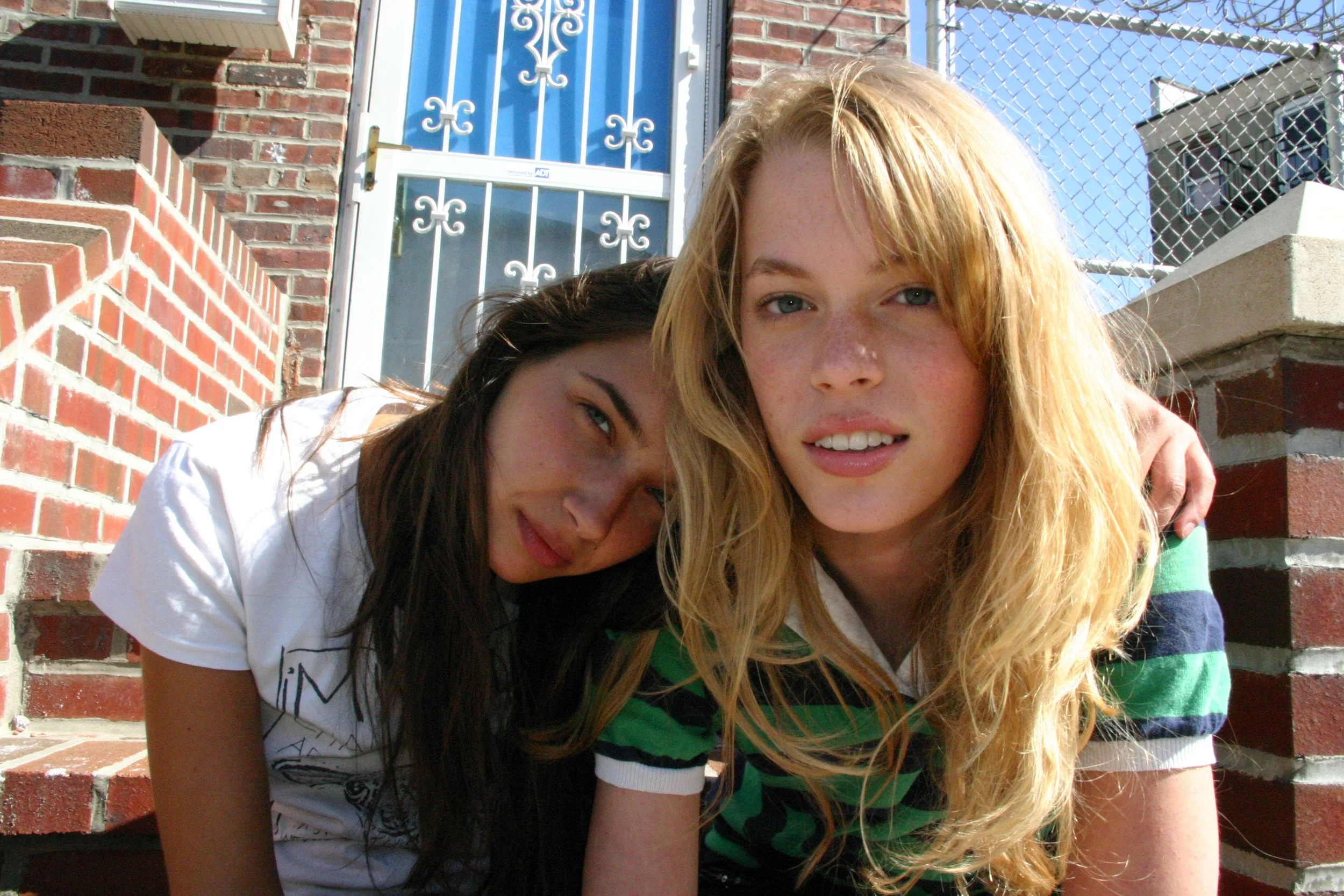 Zwei junge Frauen sitzen auf Treppen vor einer roten Backsteinmauer und einer Tür. Es ist sonnig und sie schauen in die Kamera.