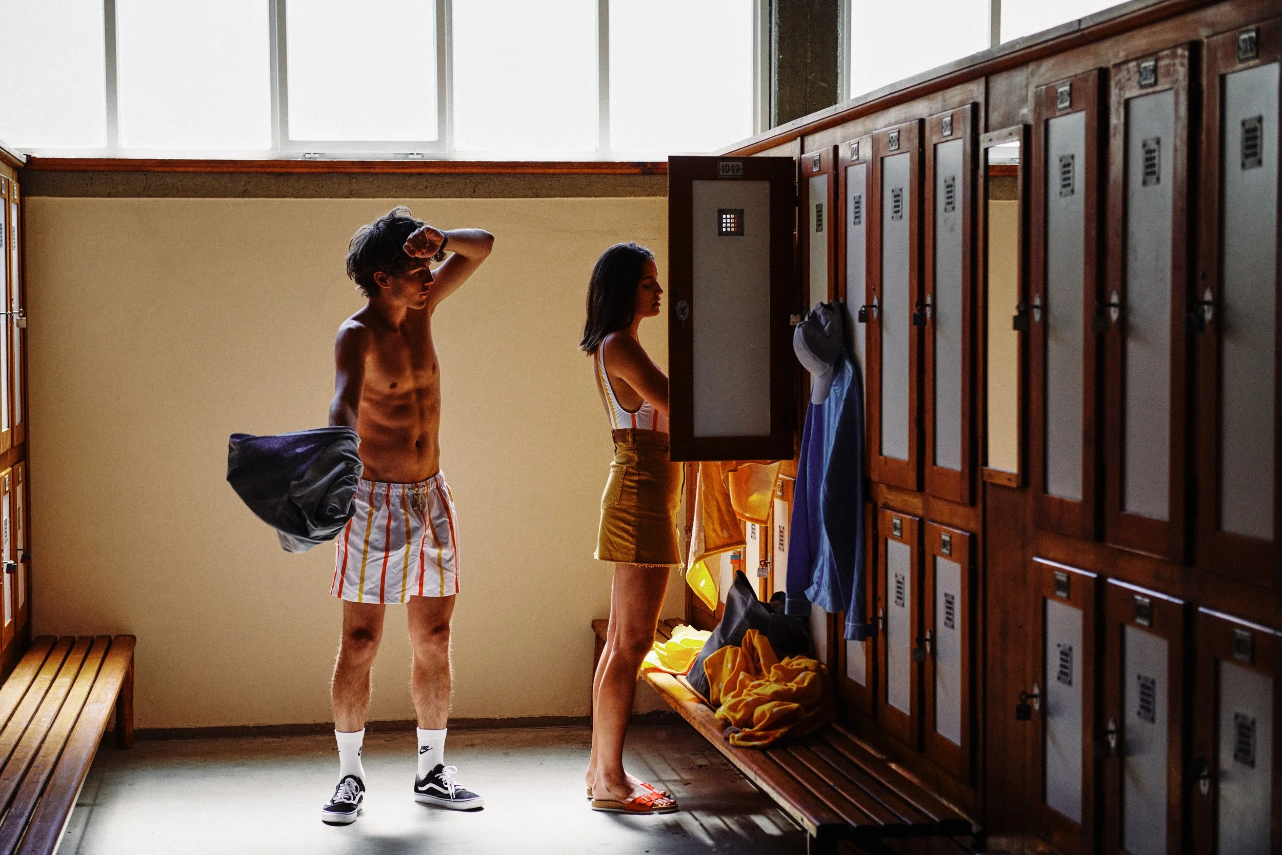 Zwei Jugendliche in einer Umkleidekabine, der junge Mann ist shirtlos und trägt Badehose, während das Mädchen vor einer Garderobe steht.