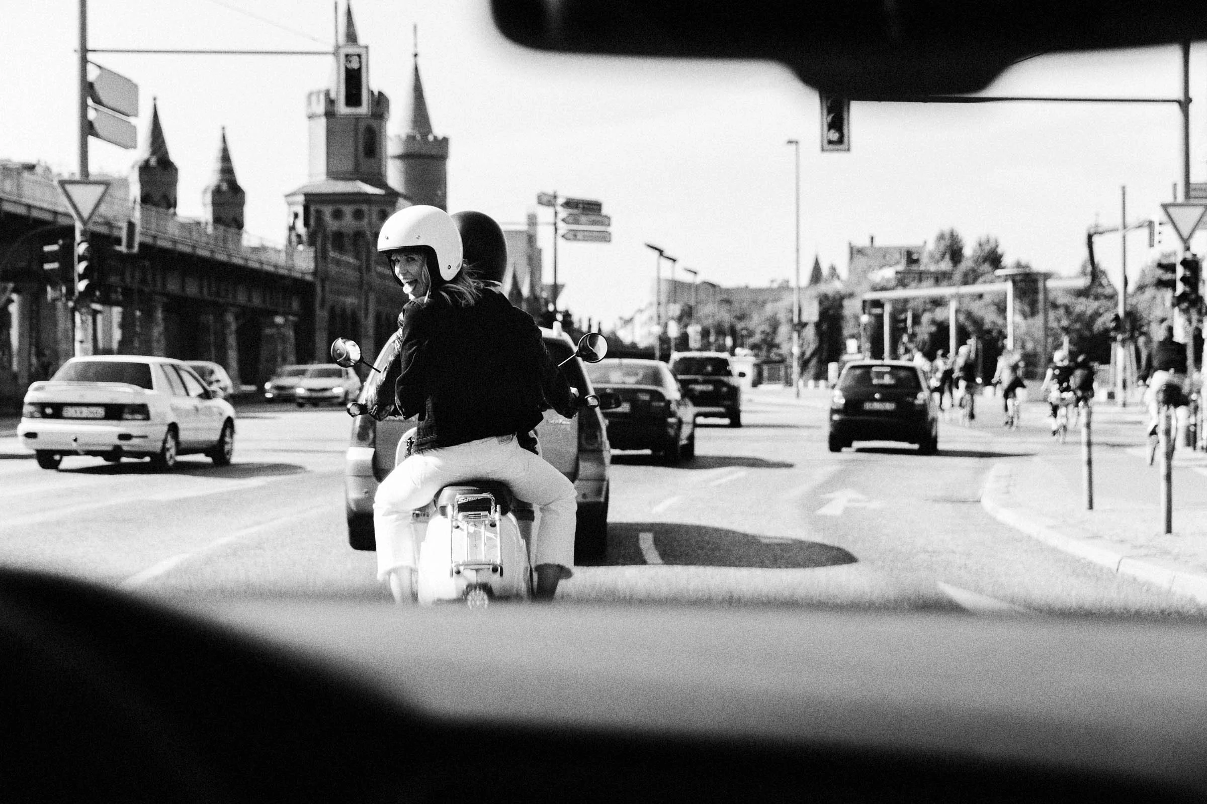 Eine schwarz-weiße Aufnahme zeigt eine junge Frau, die mit einem Helm auf einem Motorroller unterwegs ist, während sie an einer Kreuzung in einer Stadt an einer roten Ampel wartet. Im Hintergrund sind Autos, Fahrradfahrer und städtische Gebäude sicht