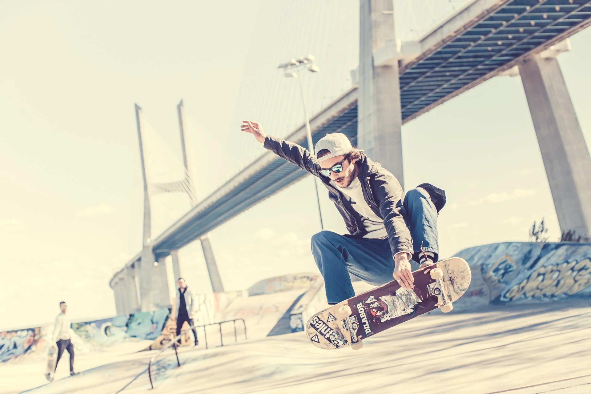 Ein junger Mann beim Skaten in einer Skateanlage unter einer Brücke, trägt eine Mütze, Sonnenbrille und eine Jacke.