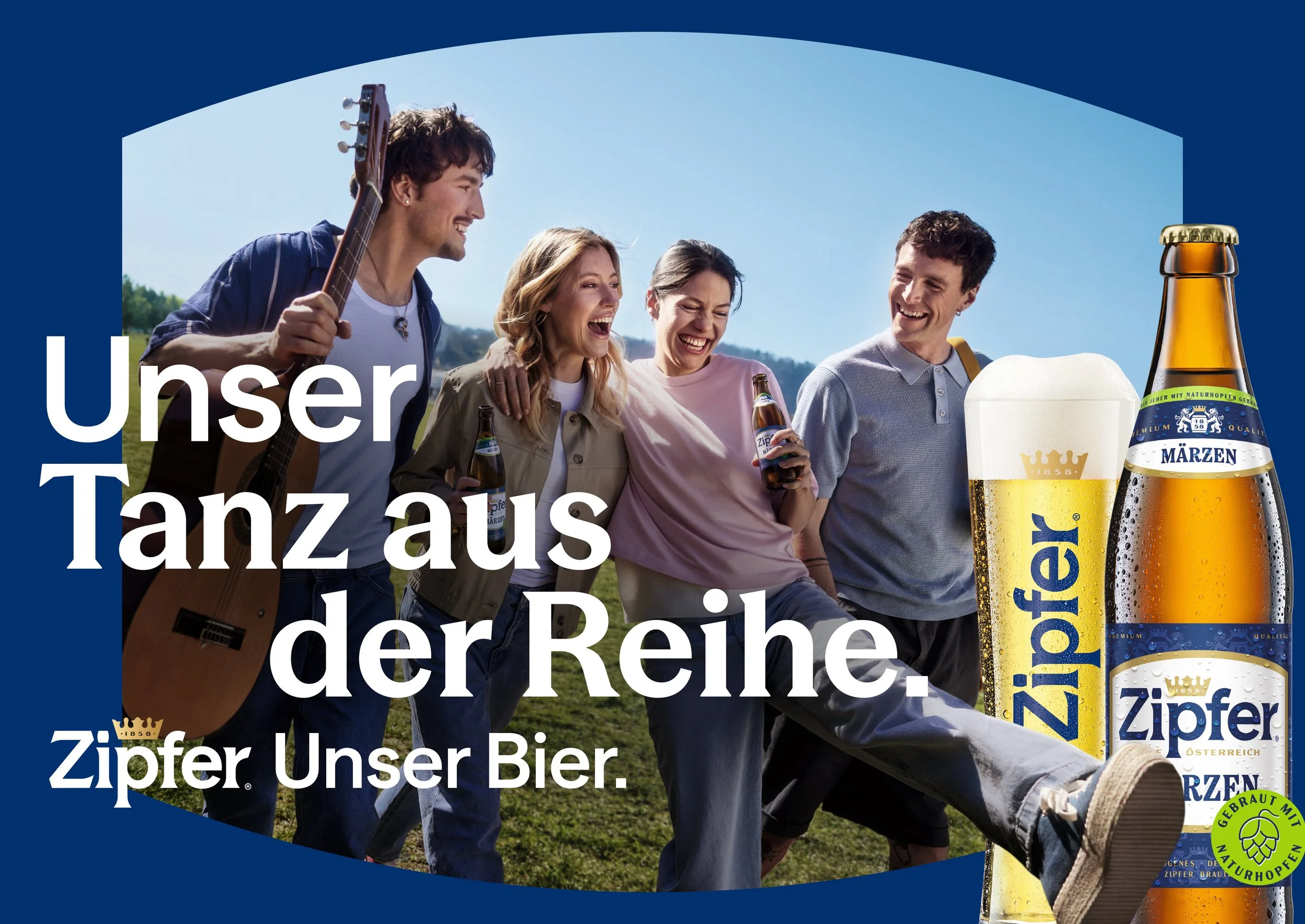 Eine Gruppe von fünf jungen Menschen, die im Freien mit Bier und einer Gitarre lachen und Spaß haben, mit Bierflaschen und einem großen Bierglas in der Nähe, vor einem blauen Himmel.