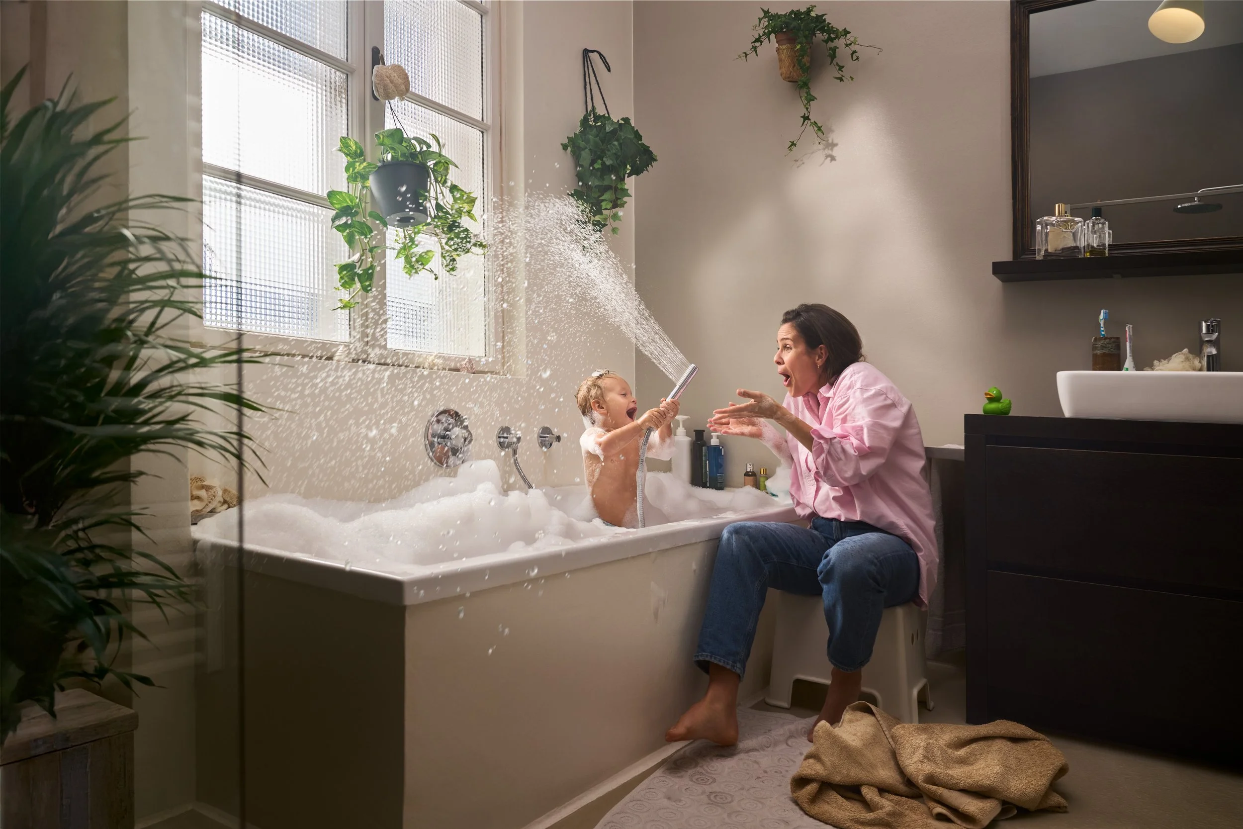Eine Frau und ein Kind im Badezimmer, das Kind spielt mit einer Gartenschlauchdüse im Badewanne, während die Frau lachend zuschaut. Das Badezimmer ist mit Pflanzen geschmückt, hat eine weiße Badewanne, Wandfliesen und einen schwarzen Waschtisch.