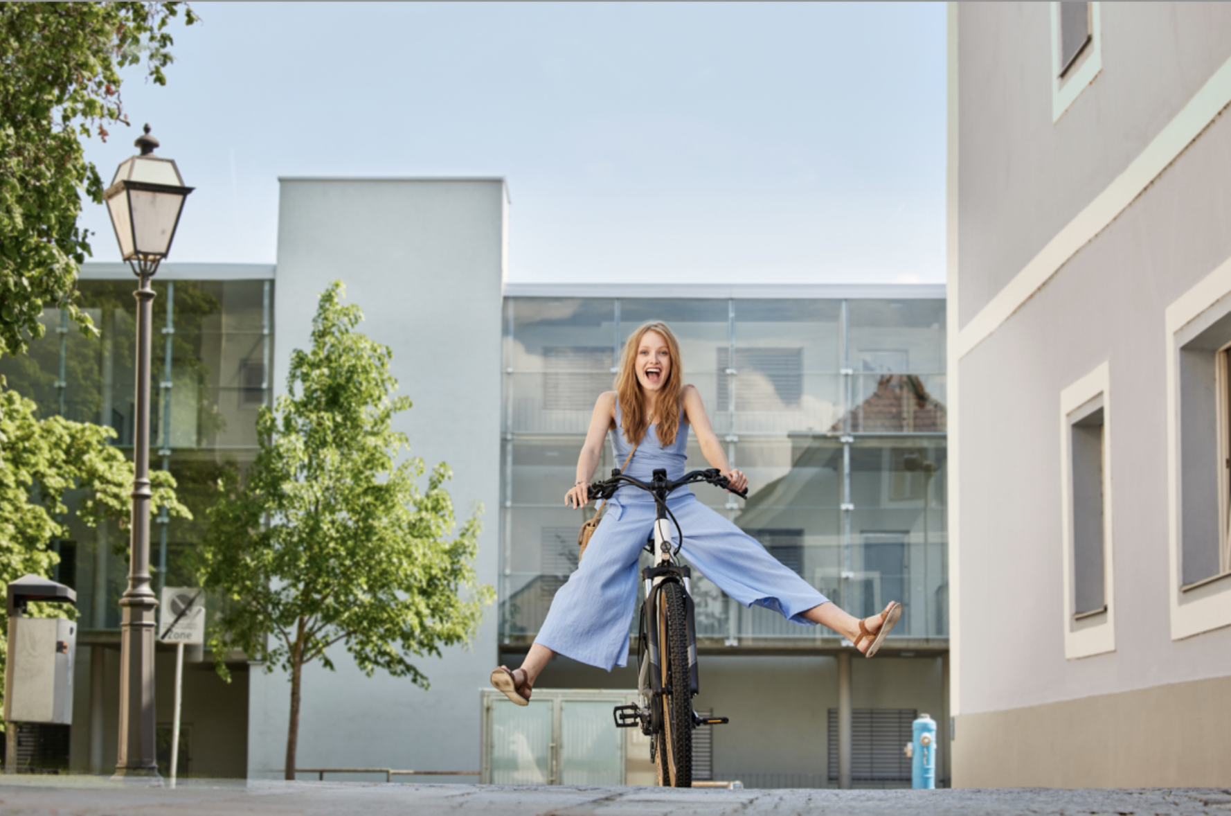 Eine lachende junge Frau in einem blauen Outfit fährt auf einem Fahrrad, hebt die Arme und hat die Beine seitlich ausgestreckt, während sie auf einer Stadtstraße unterwegs ist.