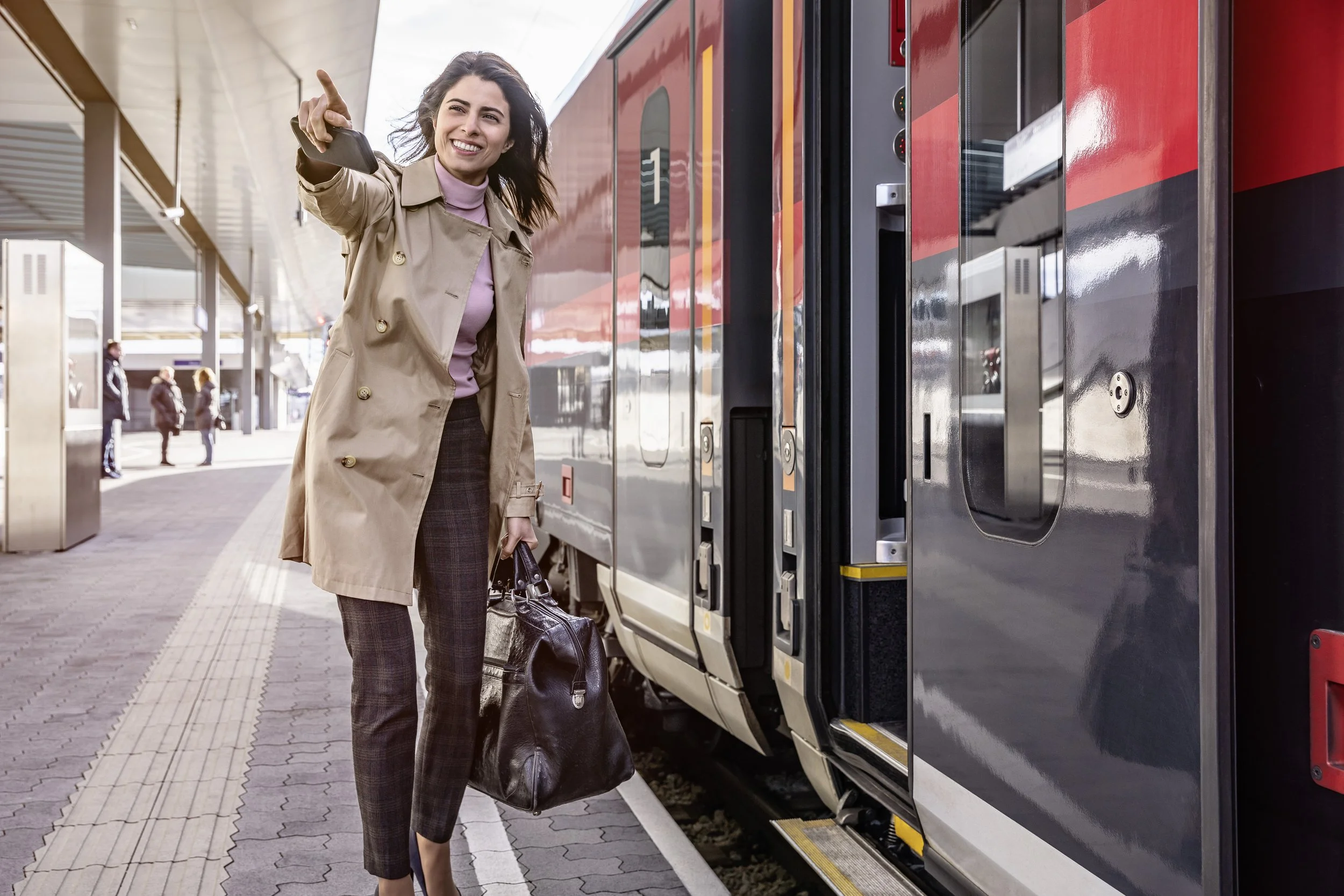 Eine lächelnde Frau in einem Trenchcoat und karierte Hose steht auf einem Bahnhof vor einem Zug, hält einen schwarzen Koffer in der Hand und macht eine Geste in Richtung Kamera.
