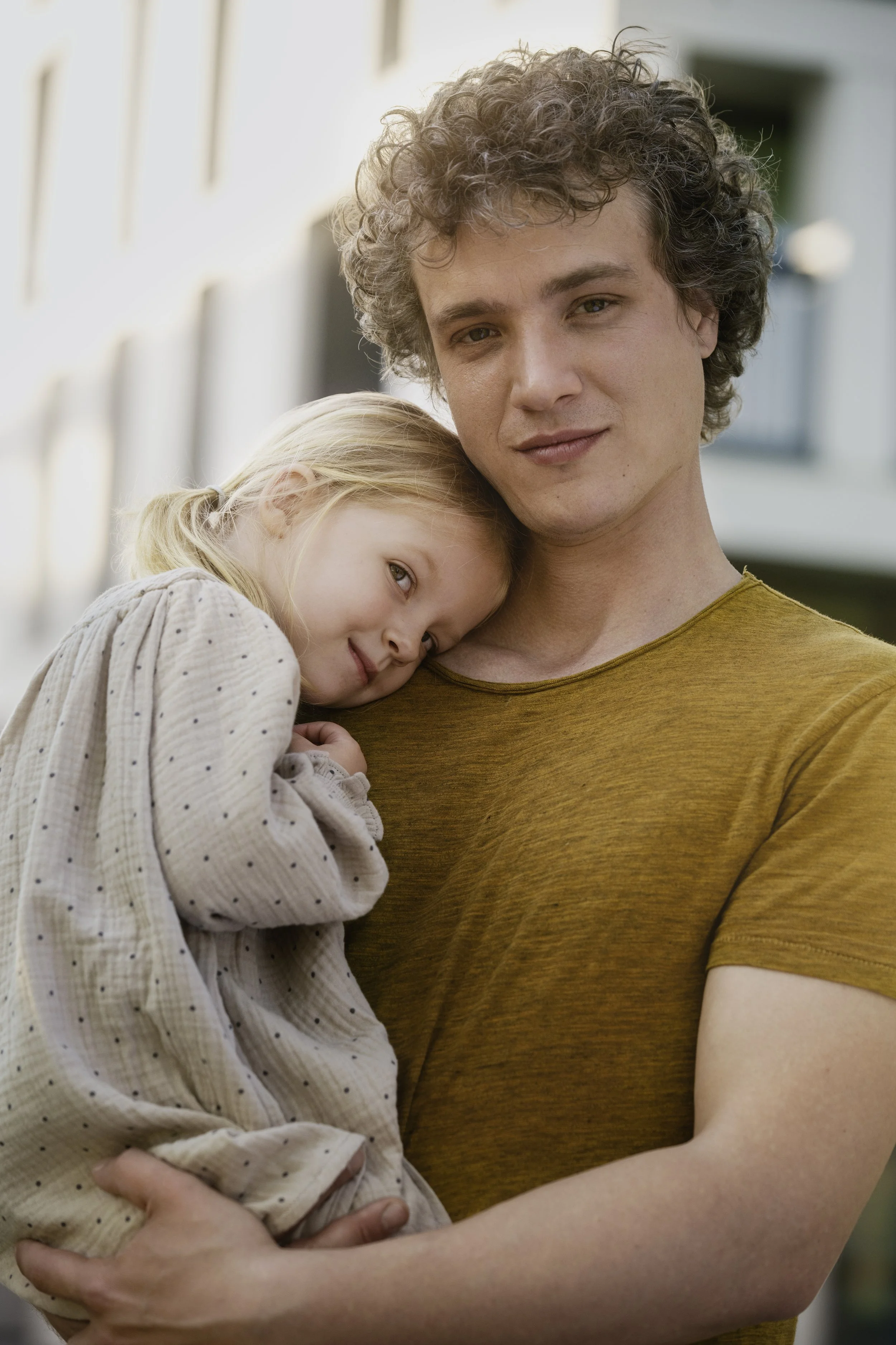 Ein junger Mann hält ein kleines Mädchen, das seinen Kopf an seine Schulter legt. Sie sind draußen vor einem modernen Gebäude, das im Hintergrund sichtbar ist.