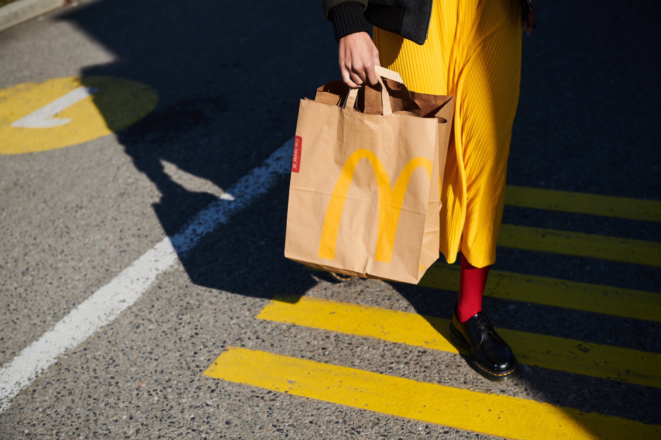 Person in gelber Kleid und roten Strümpfen, schwarze Schuhe, hält McDonald's Einkaufstasche in einer Parkgarage mit gelben Markierungen auf dem Boden.