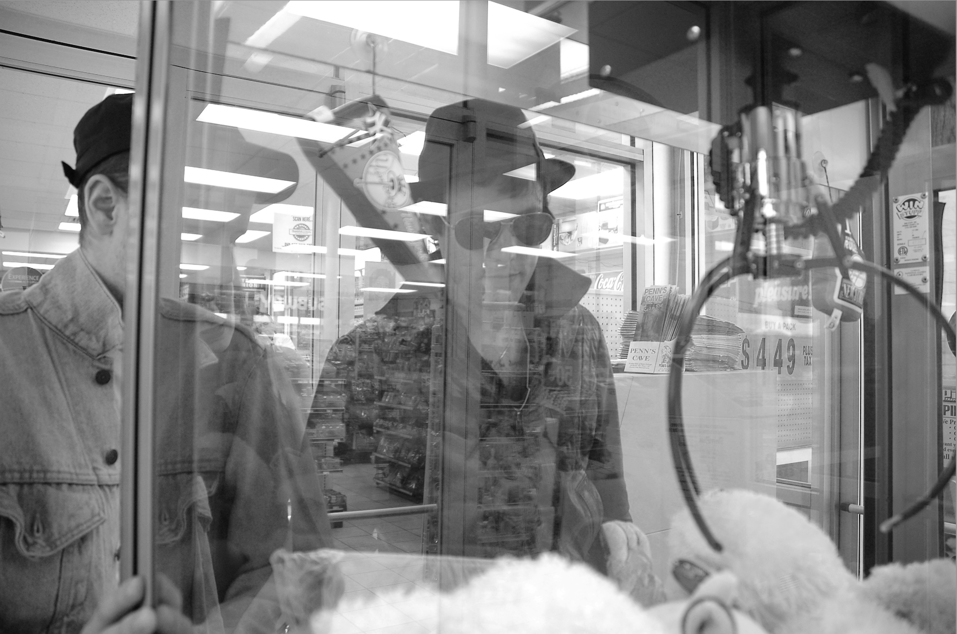 Zwei Männer schauen durch eine Glasvitrine im Supermarkt, eine trägt Sonnenbrille und einen Hut, der andere trägt eine Mütze. Eine Klammerkarte hängt an einem Spielzeug in der Vitrine.