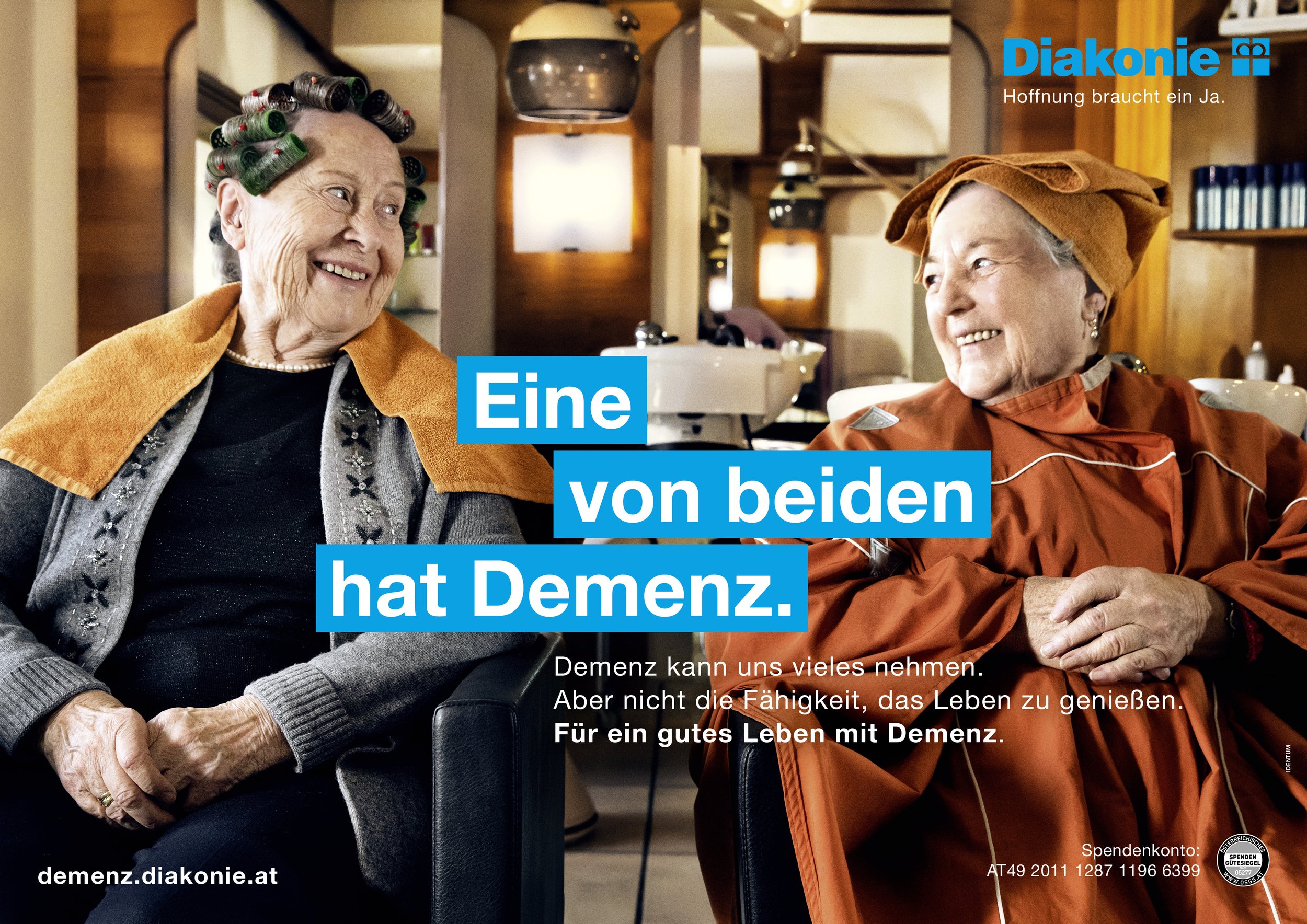 Zwei ältere Frauen sitzen in einem Salon, lachen und unterhalten sich. Beide tragen umgedrehte Hauben und bunte Umhänge, vermutlich während eines Friseurbesuchs.