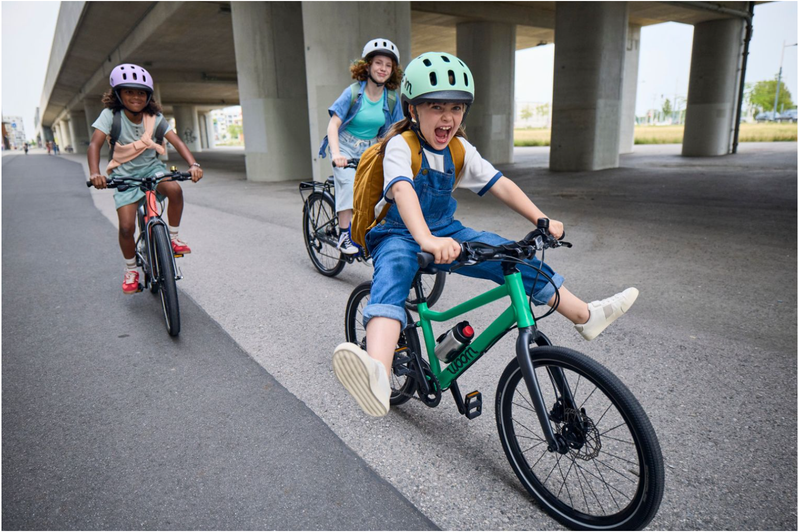 Drei Kinder fahren Fahrrad unter einer Brücke, eines lächelt, das andere schreit, alle tragen Helme und Rucksäcke.