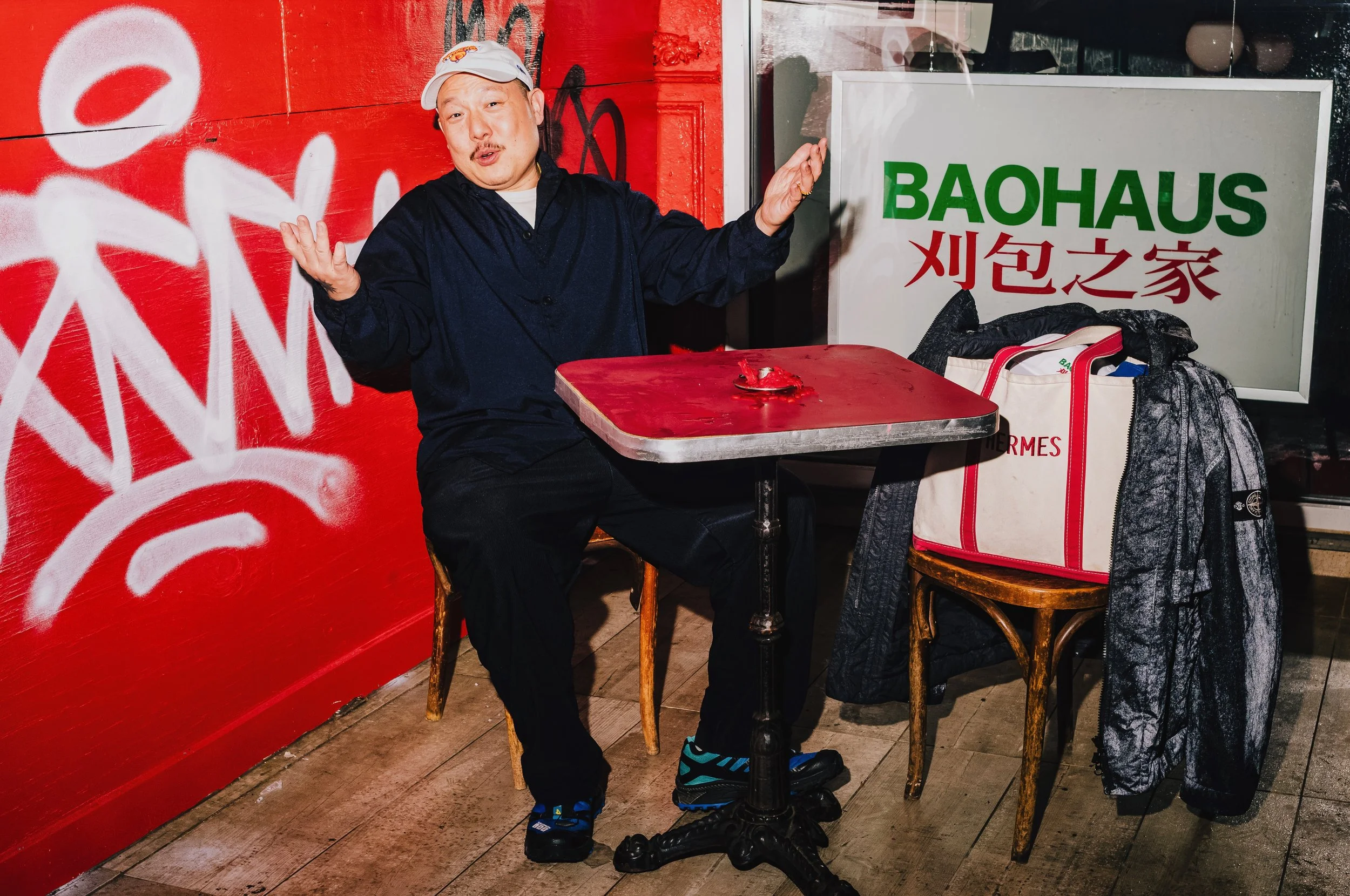 A man with light skin, wearing a white cap and black clothing, sitting at a small round table inside a restaurant. He is gesturing with his hands and has a surprised or expressive facial expression. Behind him, there is a red wall with graffiti and l