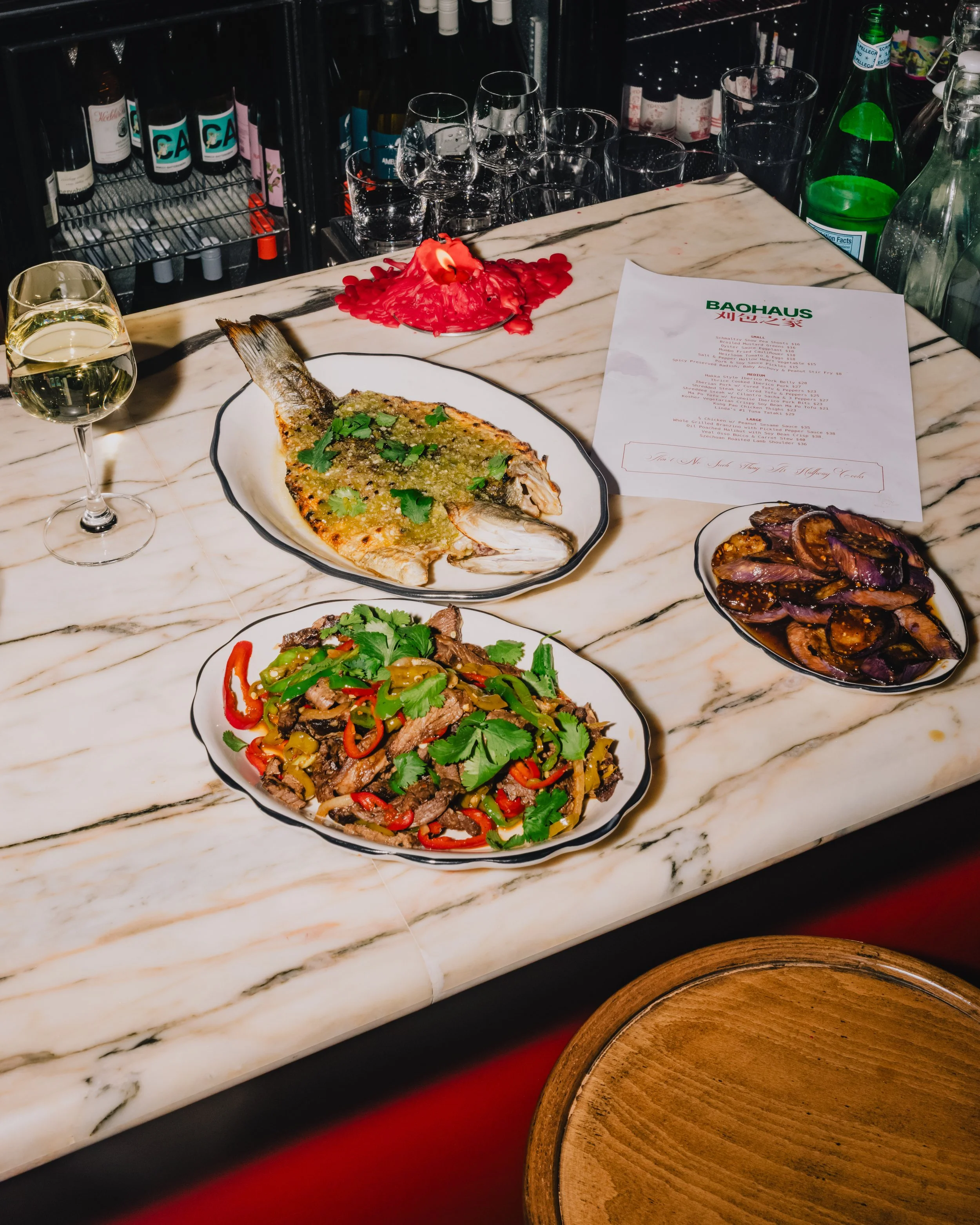 A marble countertop with a plate of fish topped with green sauce, a glass of white wine, a bowl of sliced cooked meat, a plate of stir-fried vegetables and beef, and a menu with the word 'BAUHAUS' on it.