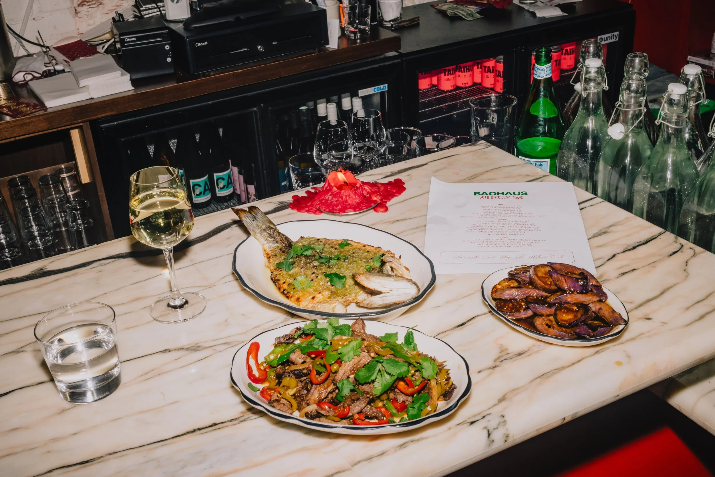 A marble countertop with various dishes including grilled fish with herbs, a colorful stir-fry with vegetables and sliced meat, and a bowl of roasted eggplant. There is a glass of white wine and a shot glass filled with water on the counter. In the b