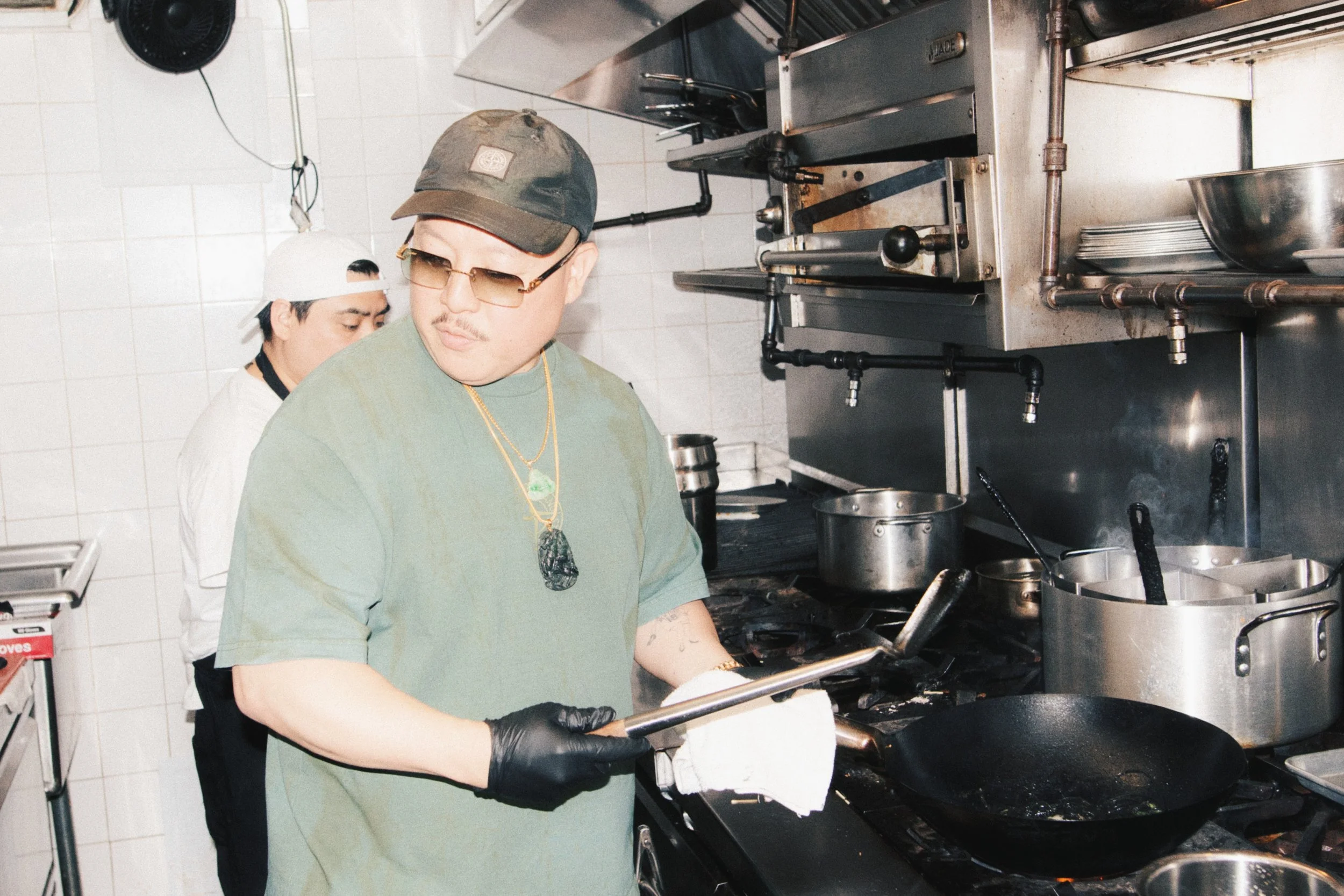 A person cooking in a commercial kitchen wearing a green T-shirt, black gloves, a hiking hat, and sunglasses. There are two men in the image, one at the front and one at the back, working at a stove with pots and kitchen equipment on stainless steel 