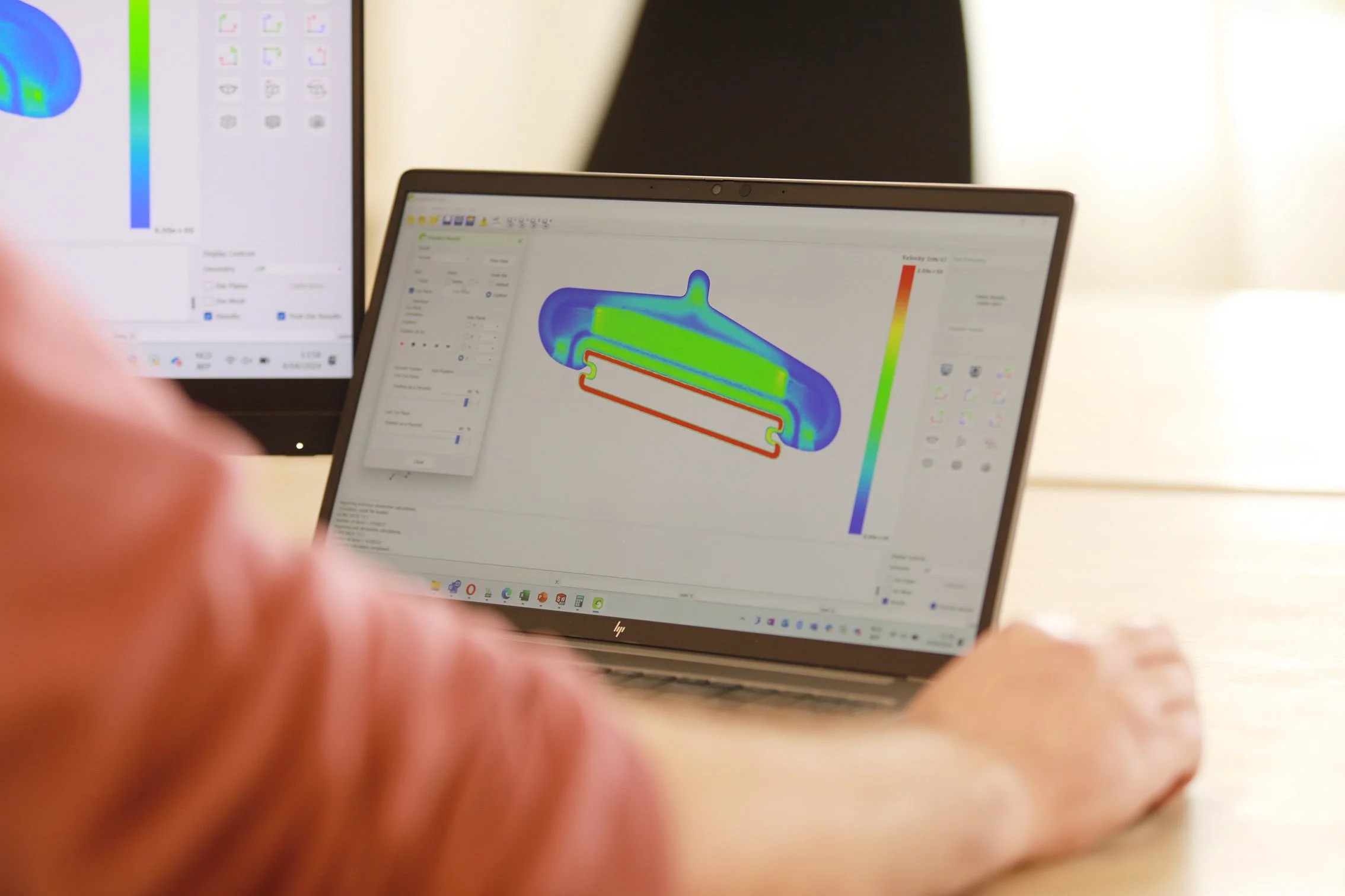 Close-up of a laptop screen showing a 3D simulation model with heatmap coloring, and another computer monitor displaying a similar simulation, possibly related to engineering or scientific research.