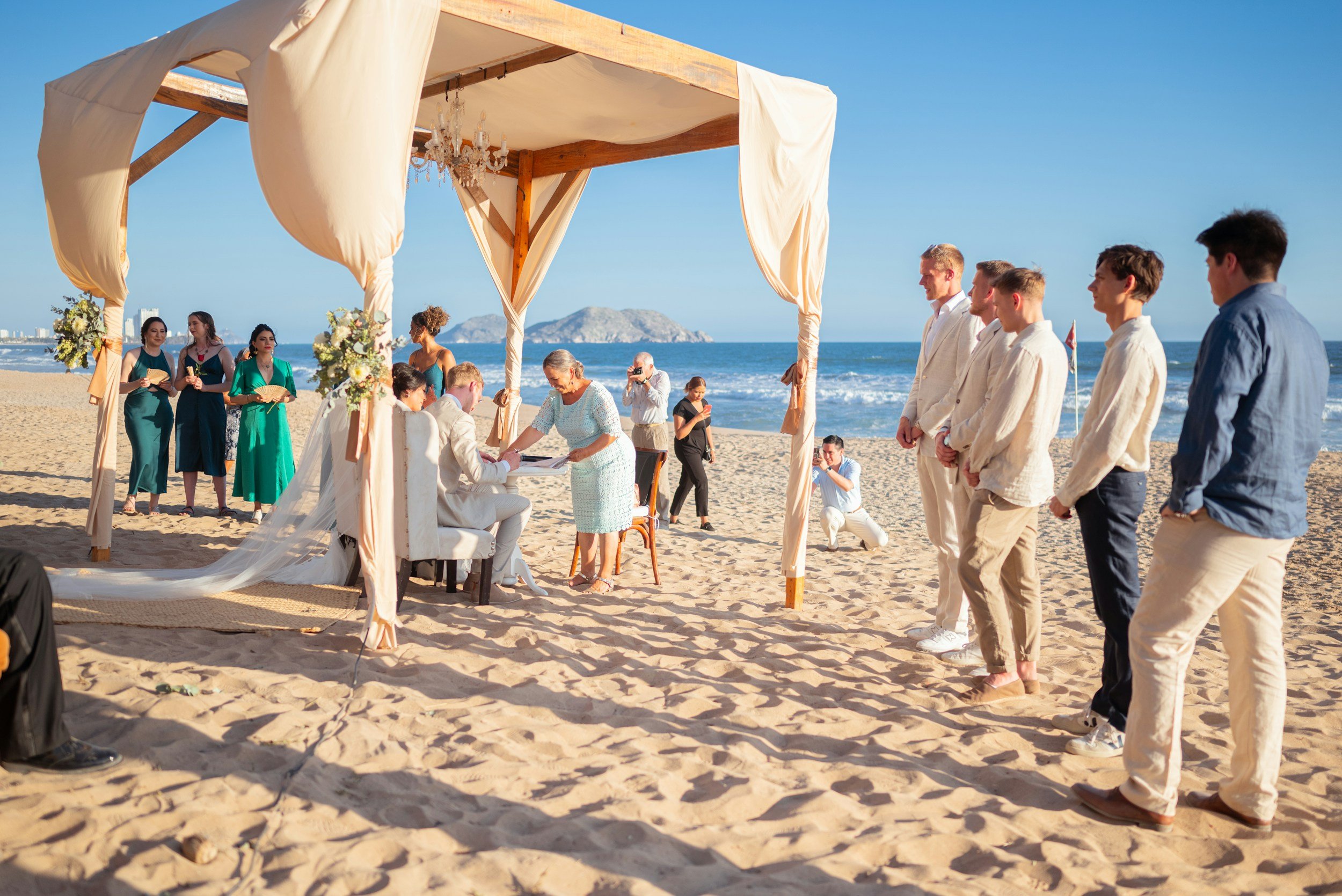 A beach wedding ceremony with a canopy, floral arrangements, and a group of people—bride and groom, officiant, bridesmaids, and groomsmen—under a bright blue sky with the ocean in the background.