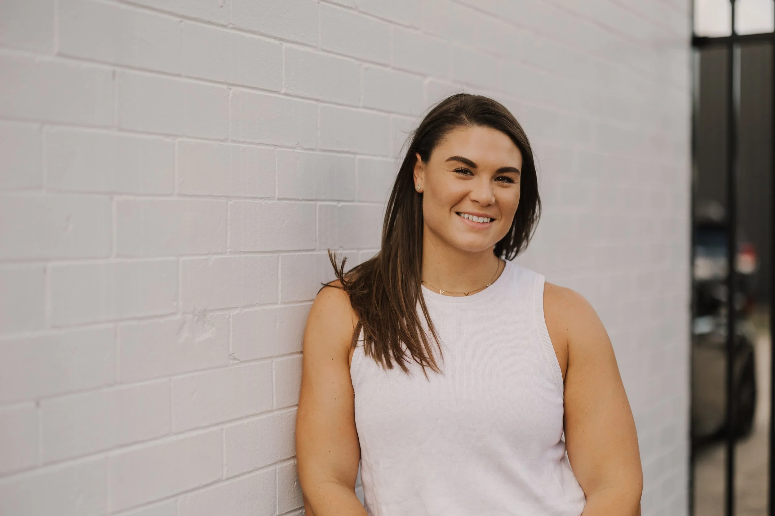 A young woman with long brown hair, wearing a white sleeveless top, smiling and leaning against a white brick wall.