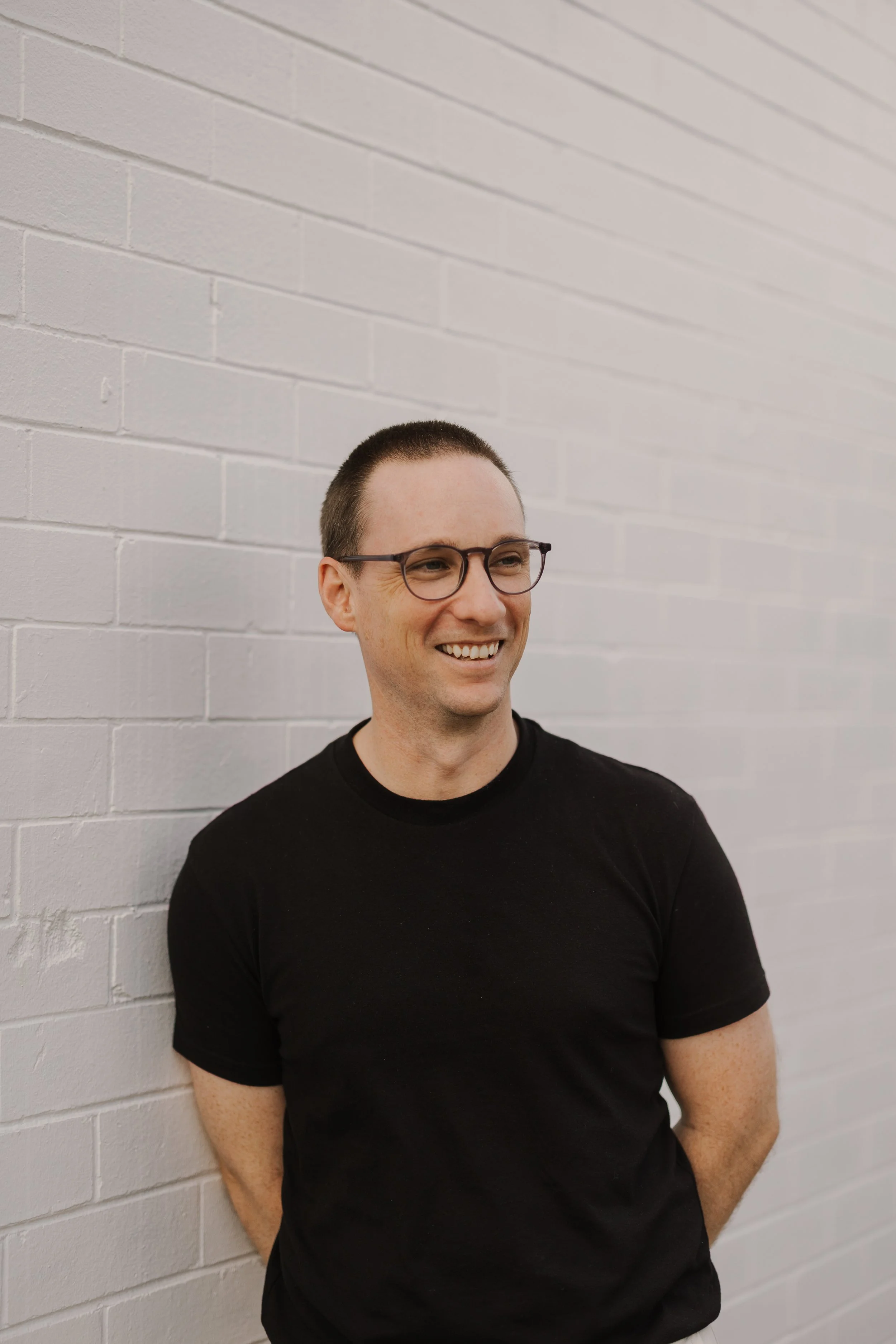 A man with short hair and glasses wearing a black t-shirt, standing against a white brick wall, smiling and looking to his left.