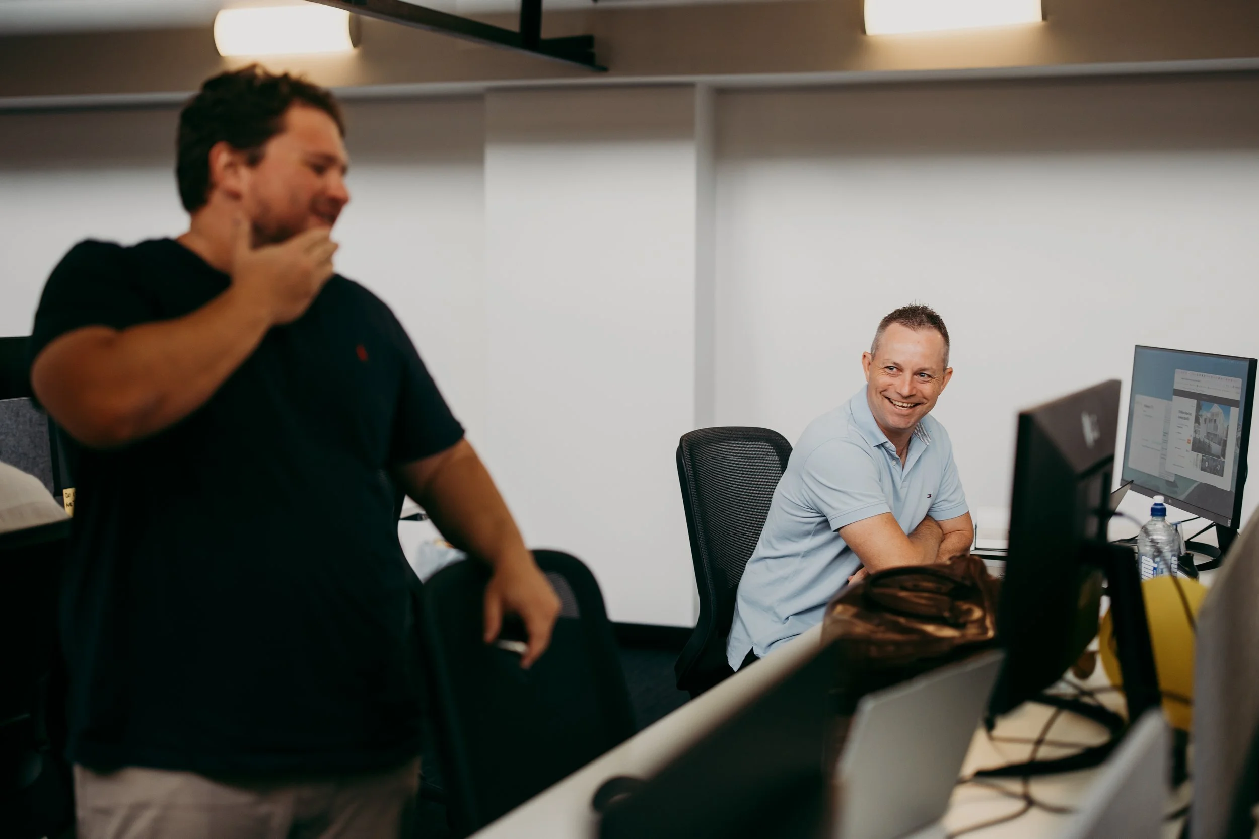 Two men working in an office, one sitting at a desk with a computer and the other standing nearby, both smiling and engaged in conversation.