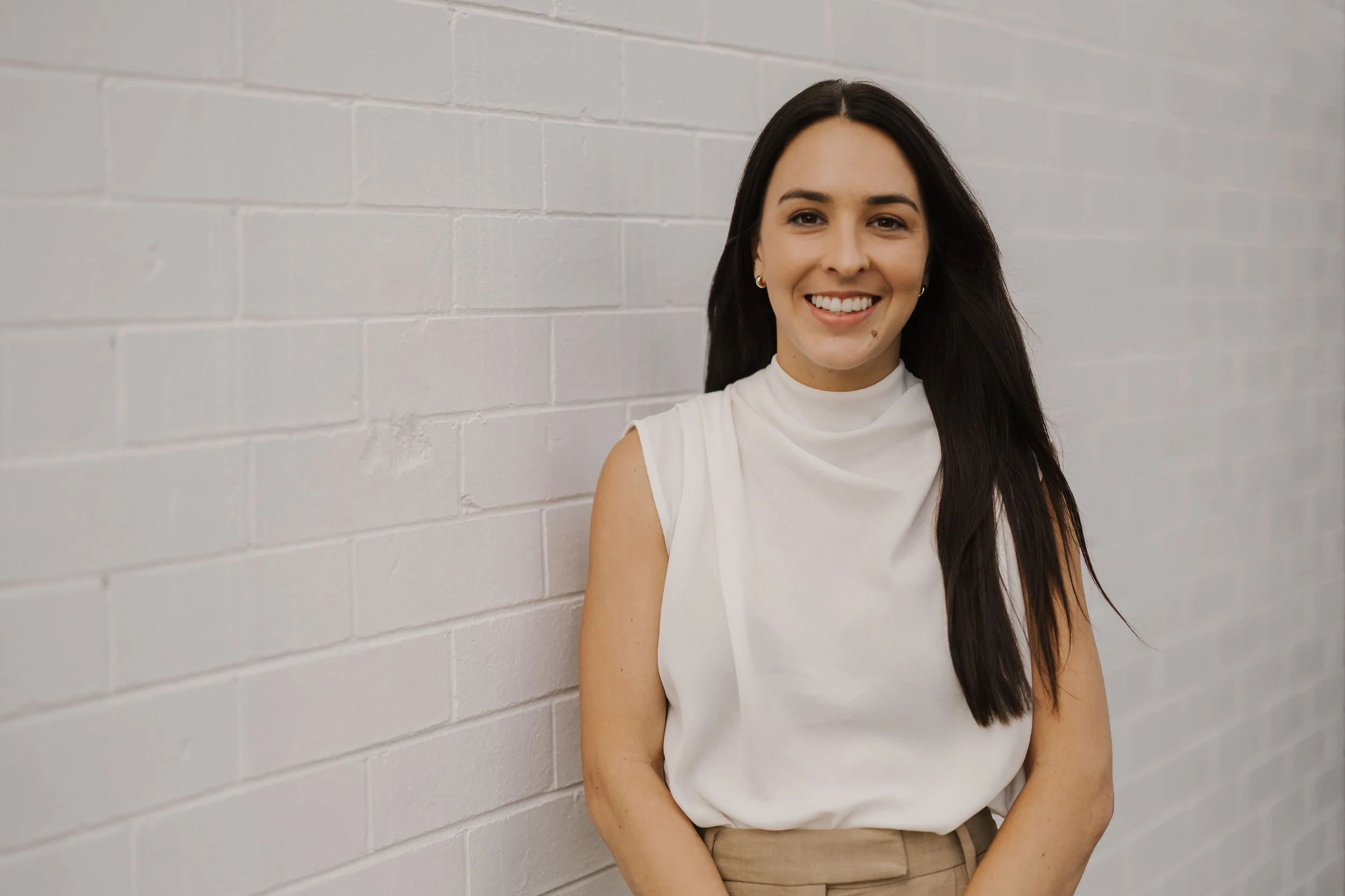 A young woman with long dark hair smiling and leaning against a white brick wall.