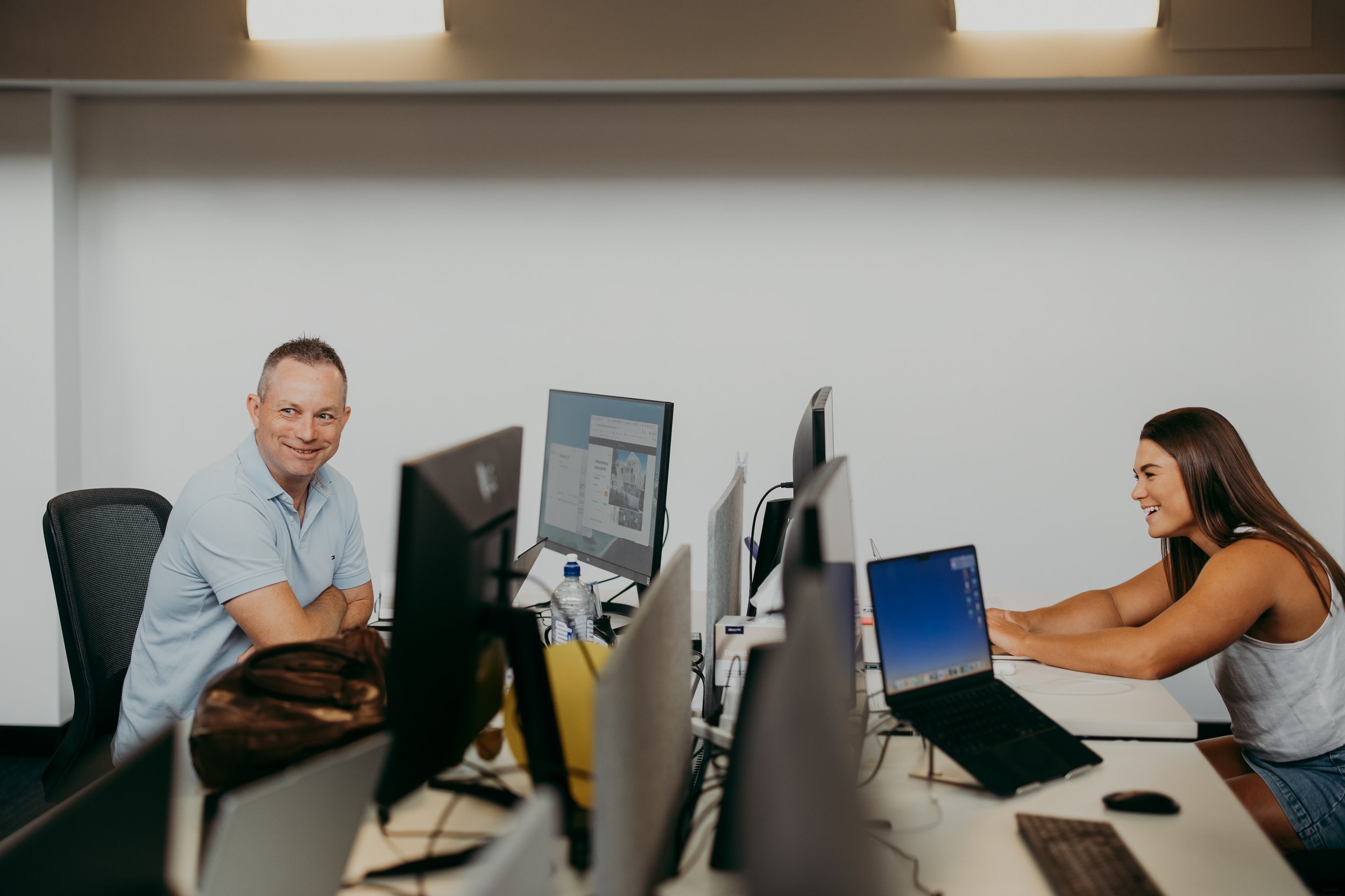 Two people sitting at desks in an office, facing computer screens, with the man smiling and the woman smiling while working.