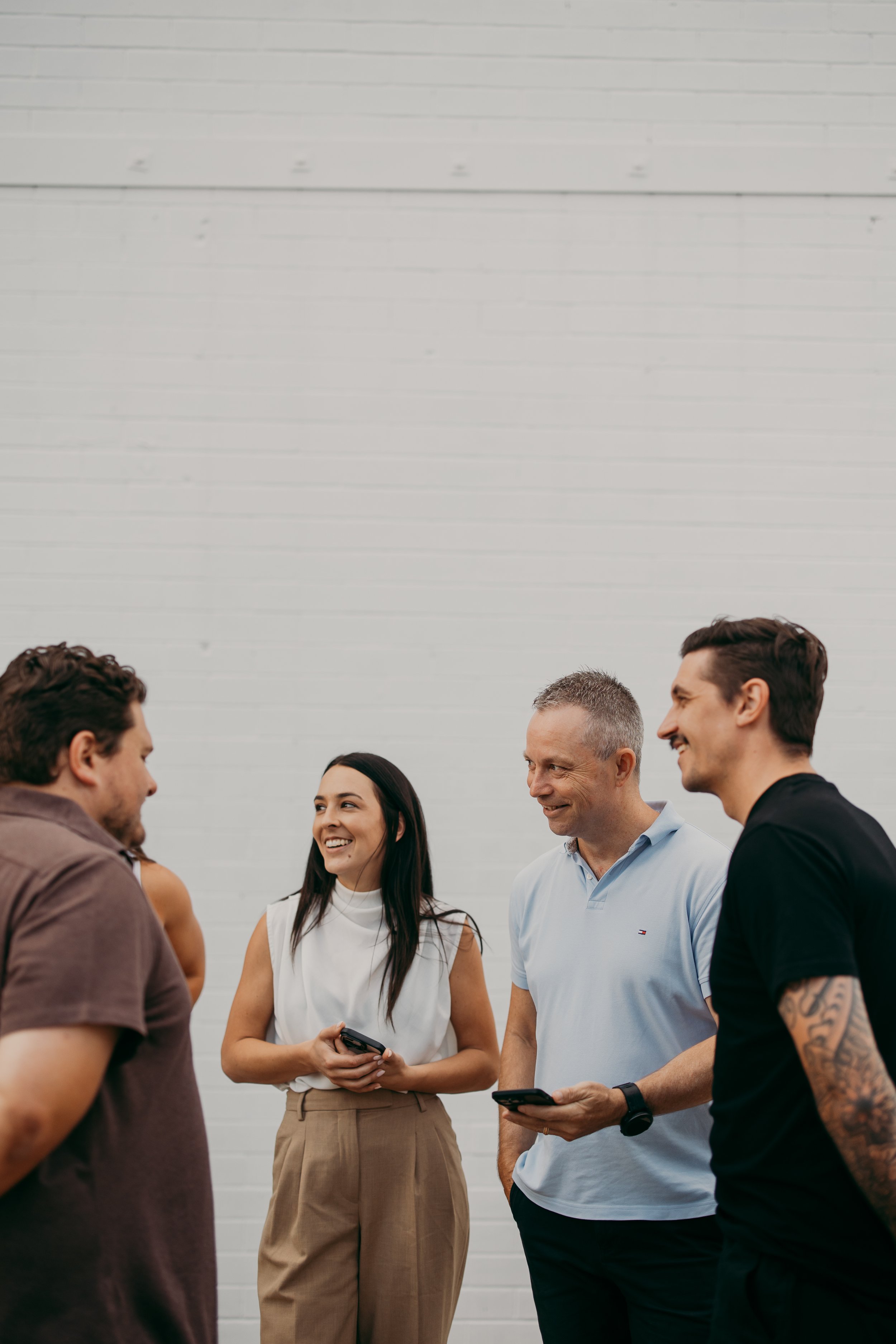 Group of five diverse adults standing and talking together, two holding smartphones, smiling, with a white brick wall background.