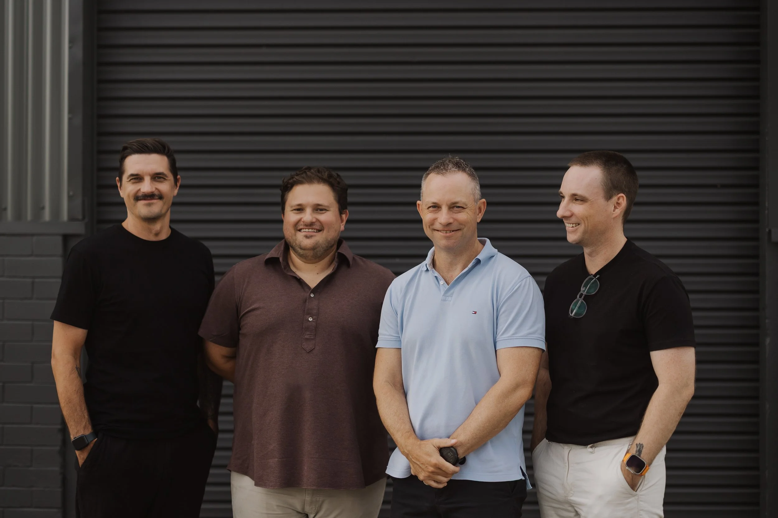 Four men standing side by side in front of a black metal garage door, smiling for the photo.
